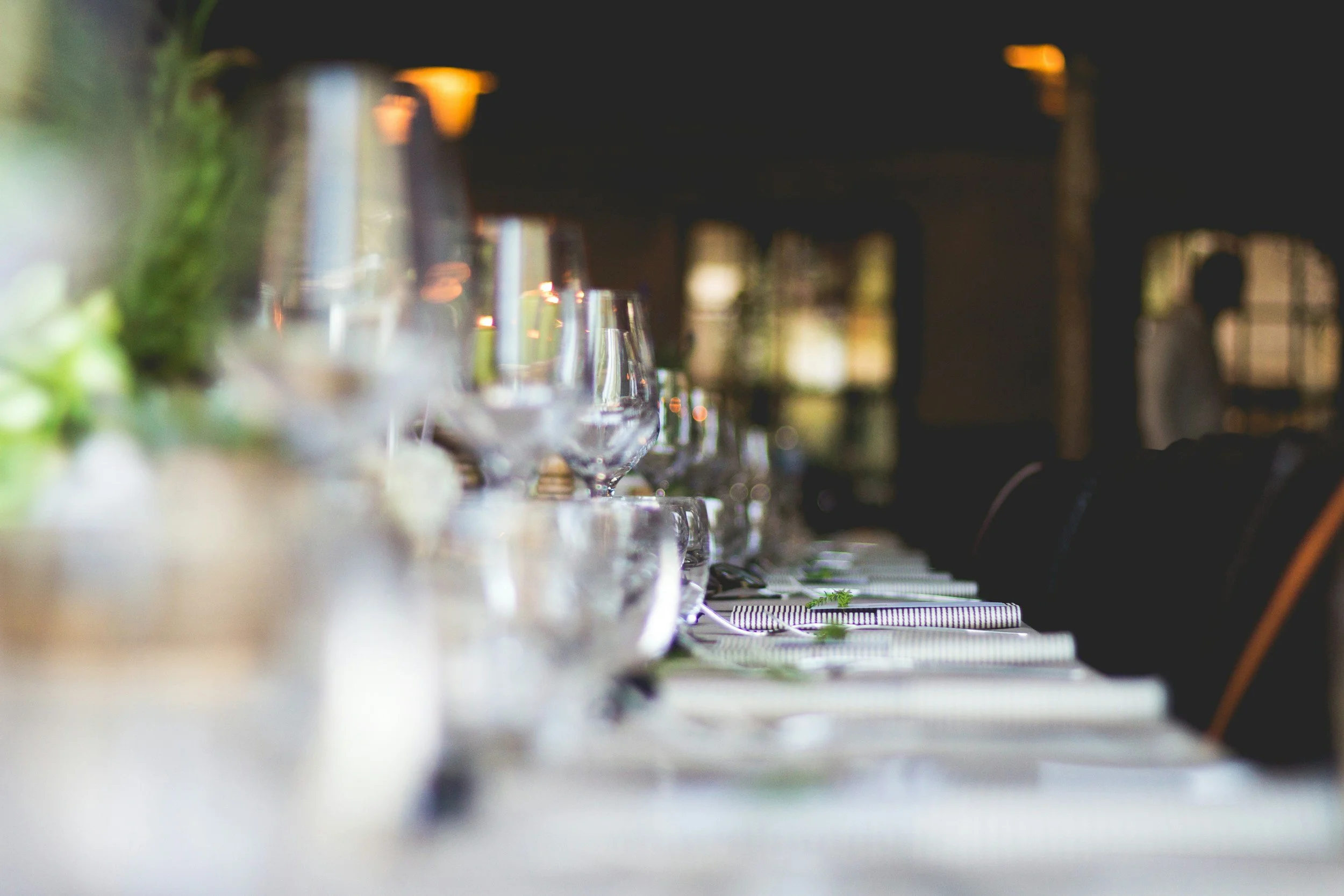 A close-up view of a long dining table set with clear wine glasses, striped napkins, and small green plants, with a blurred background of a dimly lit restaurant or event space.