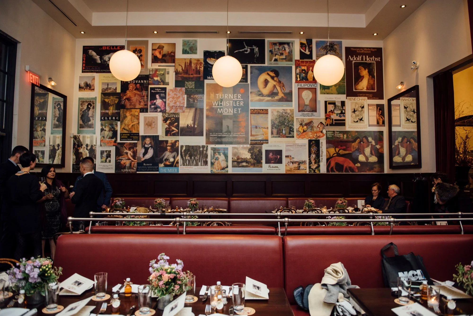 An interior view of a restaurant with a large wall decorated with numerous art posters, and three globular pendant lights hanging from the ceiling. There are groups of people socializing, and a table set with flowers, menus, and glasses in the foreground.