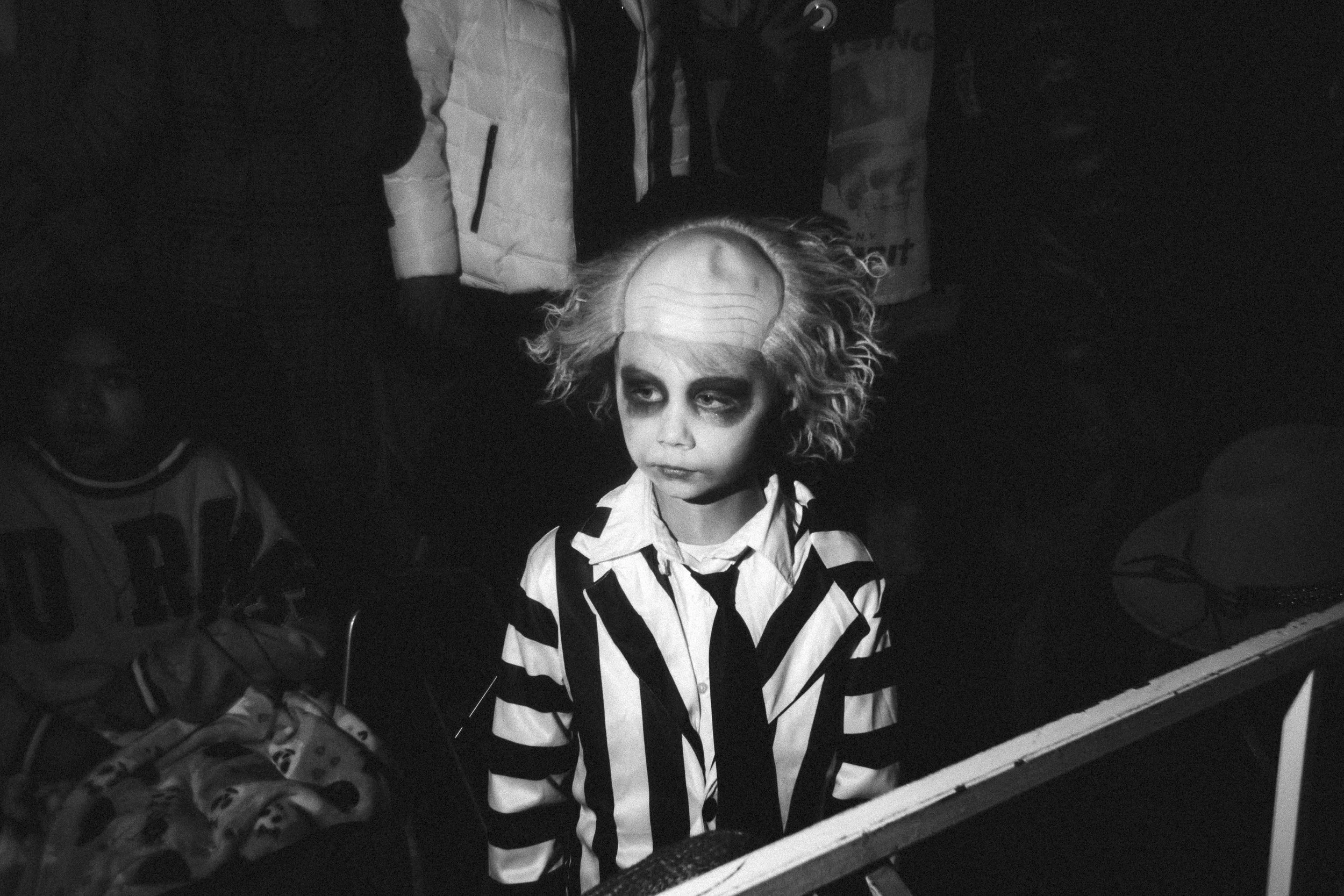 A young girl with curly hair wearing a black and white striped sweater, looking slightly to the side with a serious expression. She appears to be at a costume event or photo shoot, with a dark background and some indistinct objects around her.