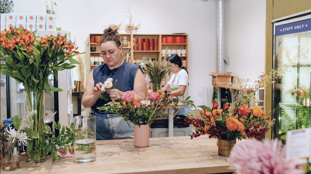 Two women arranging flowers in a shop filled with colorful flower arrangements and vases, with shelves of supplies in the background.