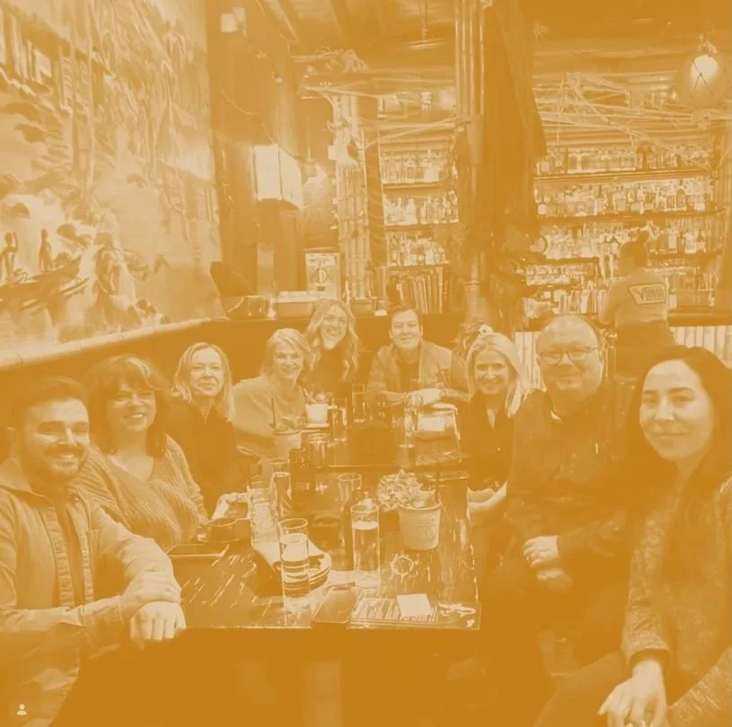 A group of ten people sitting around a table in a restaurant or pub, smiling at the camera with drinks in front of them.