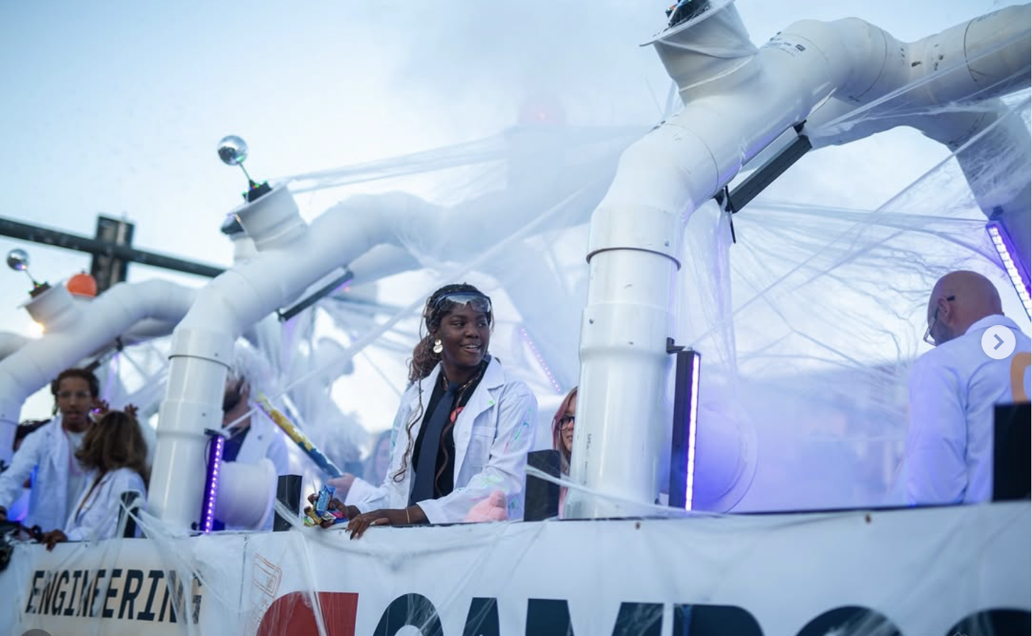 Women in lab coats at a scientific event with large robotic arms and a white canopy overhead.