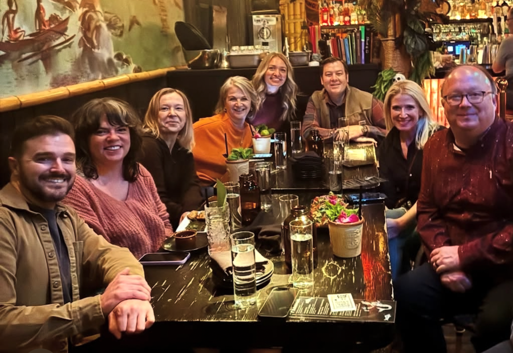 Group of eight people sitting around a restaurant table, smiling, with drinks and small plants on the table, and a colorful mural and bar in the background.