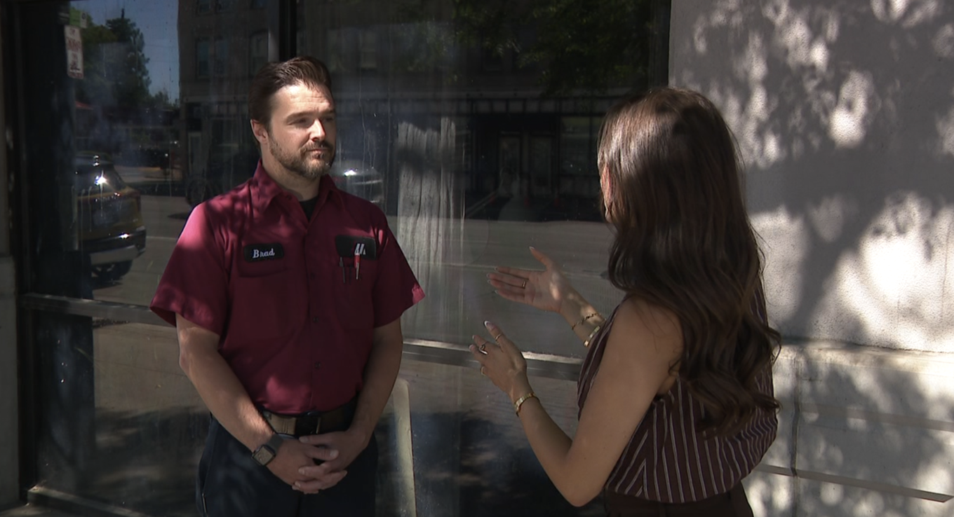 A man in a maroon uniform with a name tag that says 'Brad' is standing outside in front of a glass window, talking to a woman with long brown hair wearing a sleeveless striped top, on a sunny day with shadows of trees on the wall behind them.
