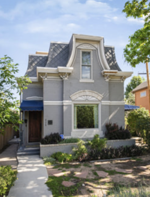 A small gray Victorian-style house with a curved roof, front window, and front door, surrounded by a garden and trees.