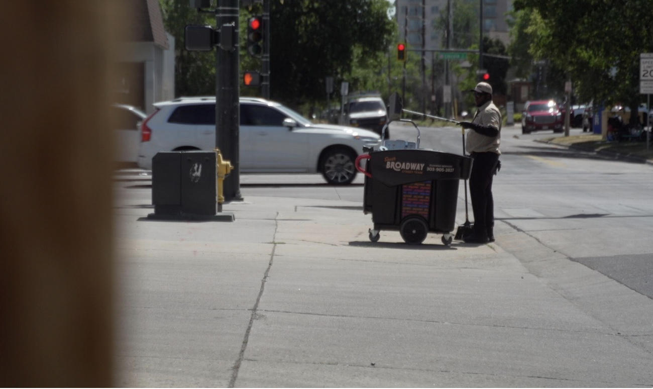 A street corner with a man dressed in a beige shirt and dark pants standing next to a large black cart and talking on a phone. Cars are visible in the background at a red traffic light, and trees line the street.
