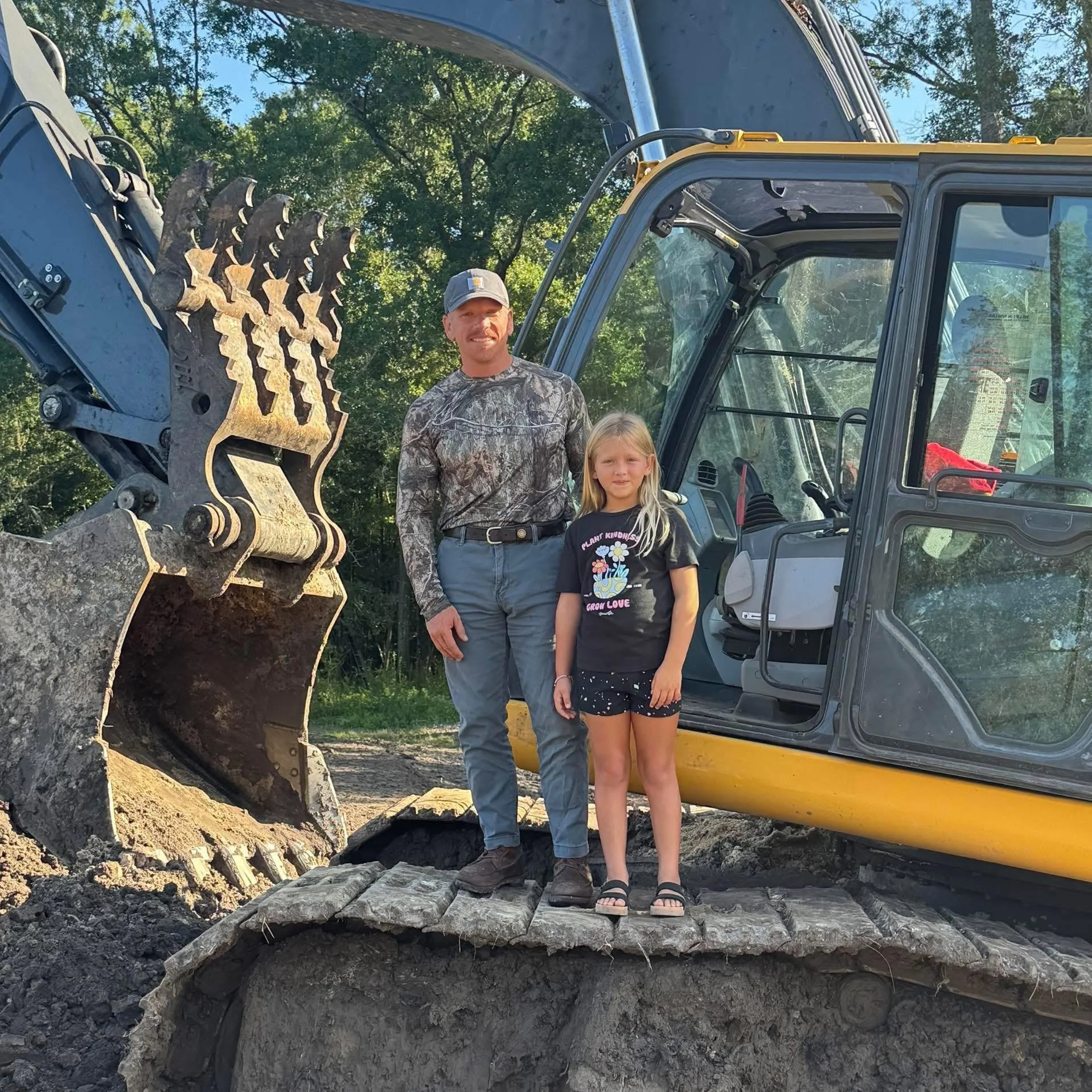 A man and a young girl standing in front of a large piece of construction equipment, possibly an excavator, outdoors with trees in the background. The man is wearing a camouflage shirt, gray pants, and a gray cap, and the girl is wearing a black T-shirt with colorful flowers and black shorts. They are standing on uneven ground with pieces of metal and dirt around.