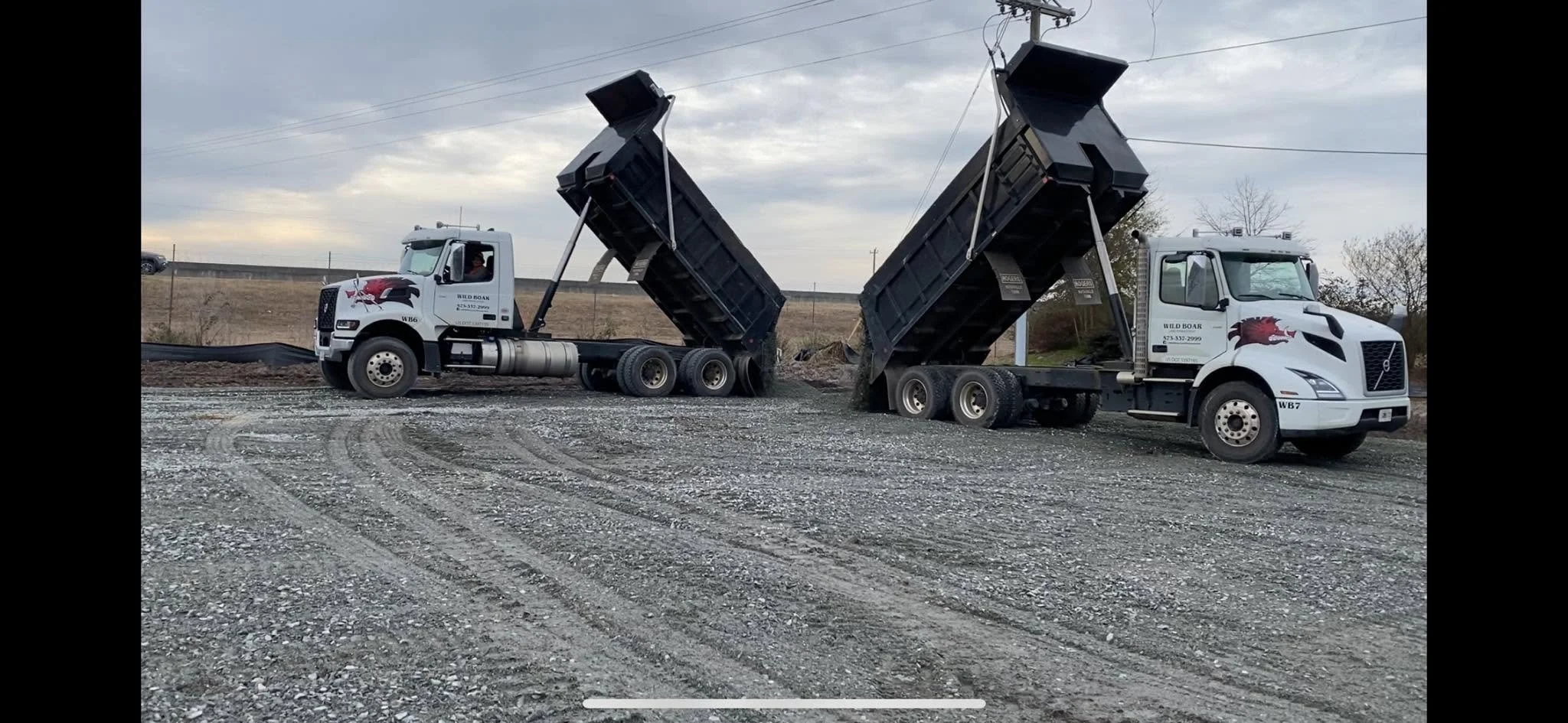 Two dump trucks with their beds raised, unloading gravel on a construction site under a cloudy sky.