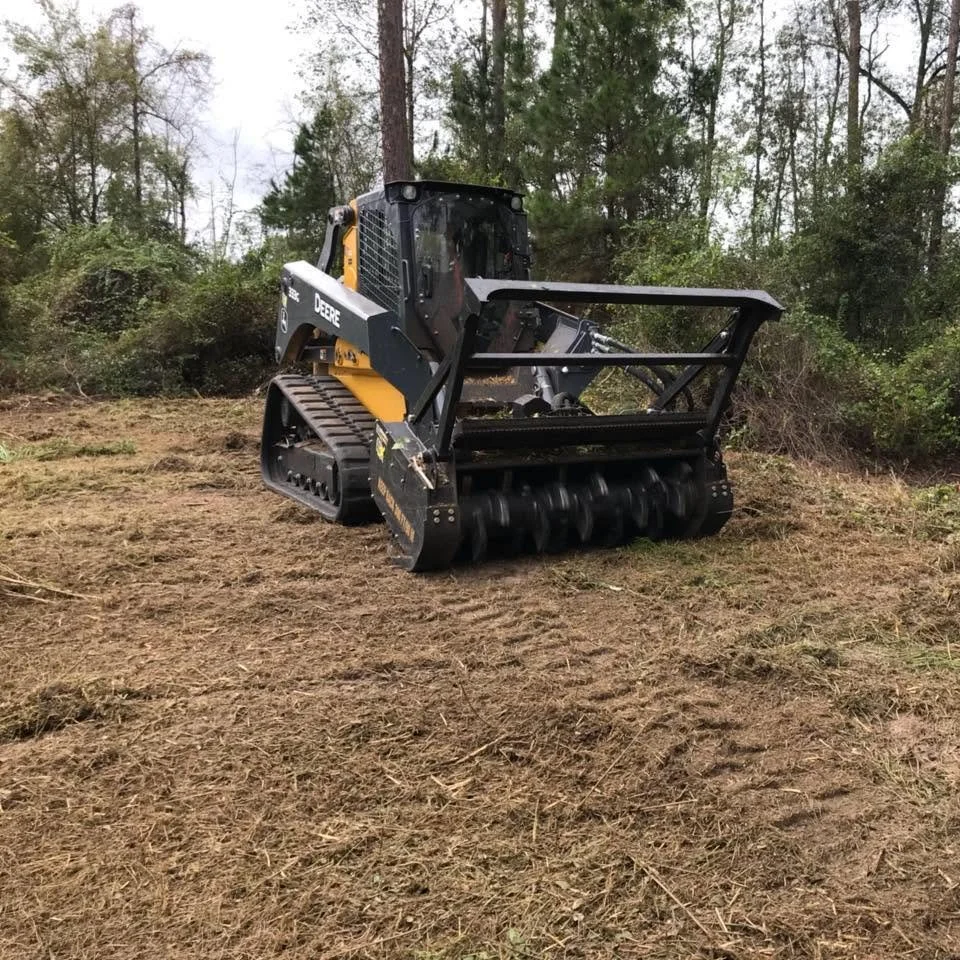 A small, black and yellow John Deere skid steer loader with a roller attachment working on a dirt clearing site surrounded by trees.