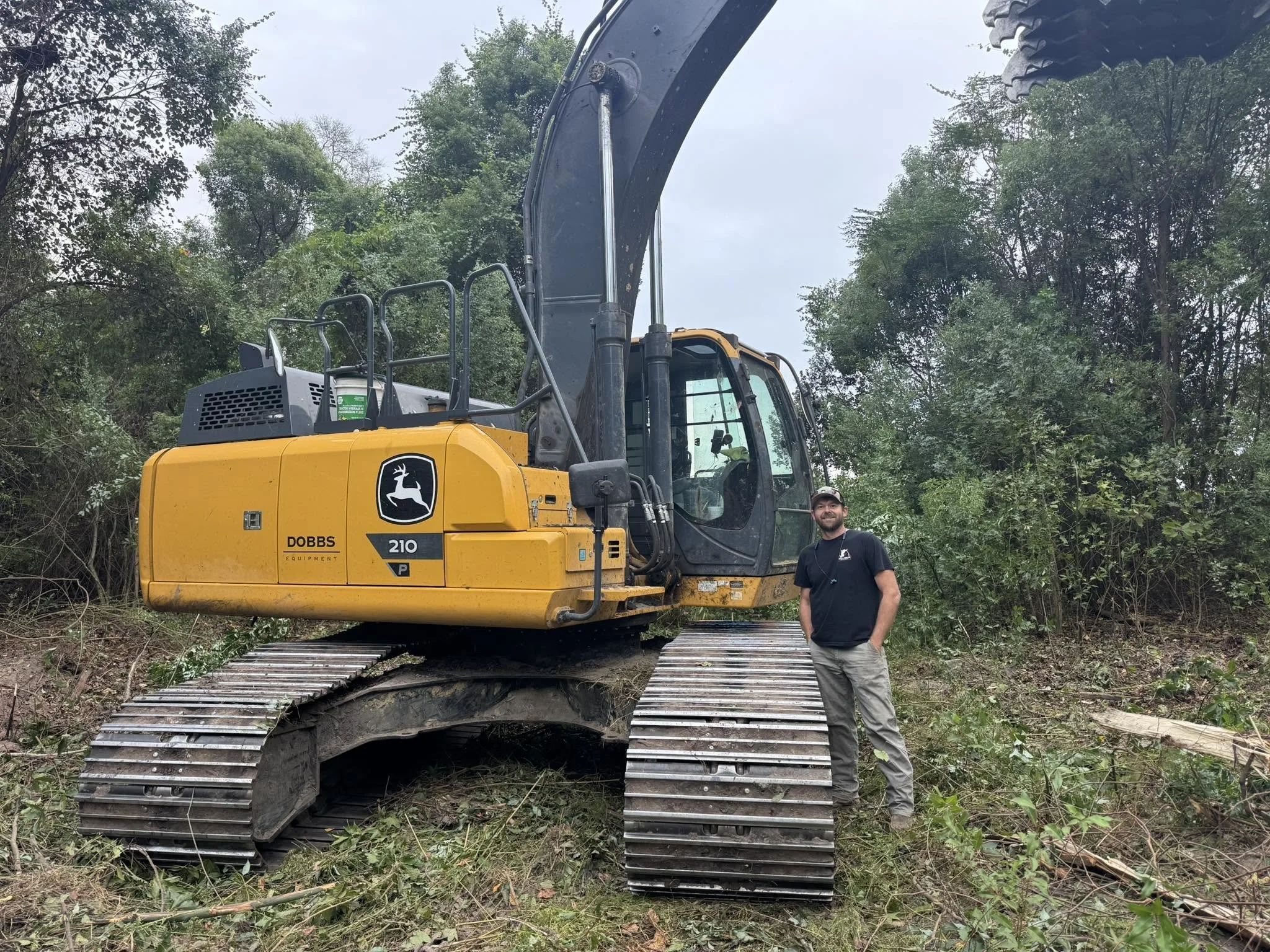 A man standing next to a large yellow John Deere excavator with a black arm in a wooded area.