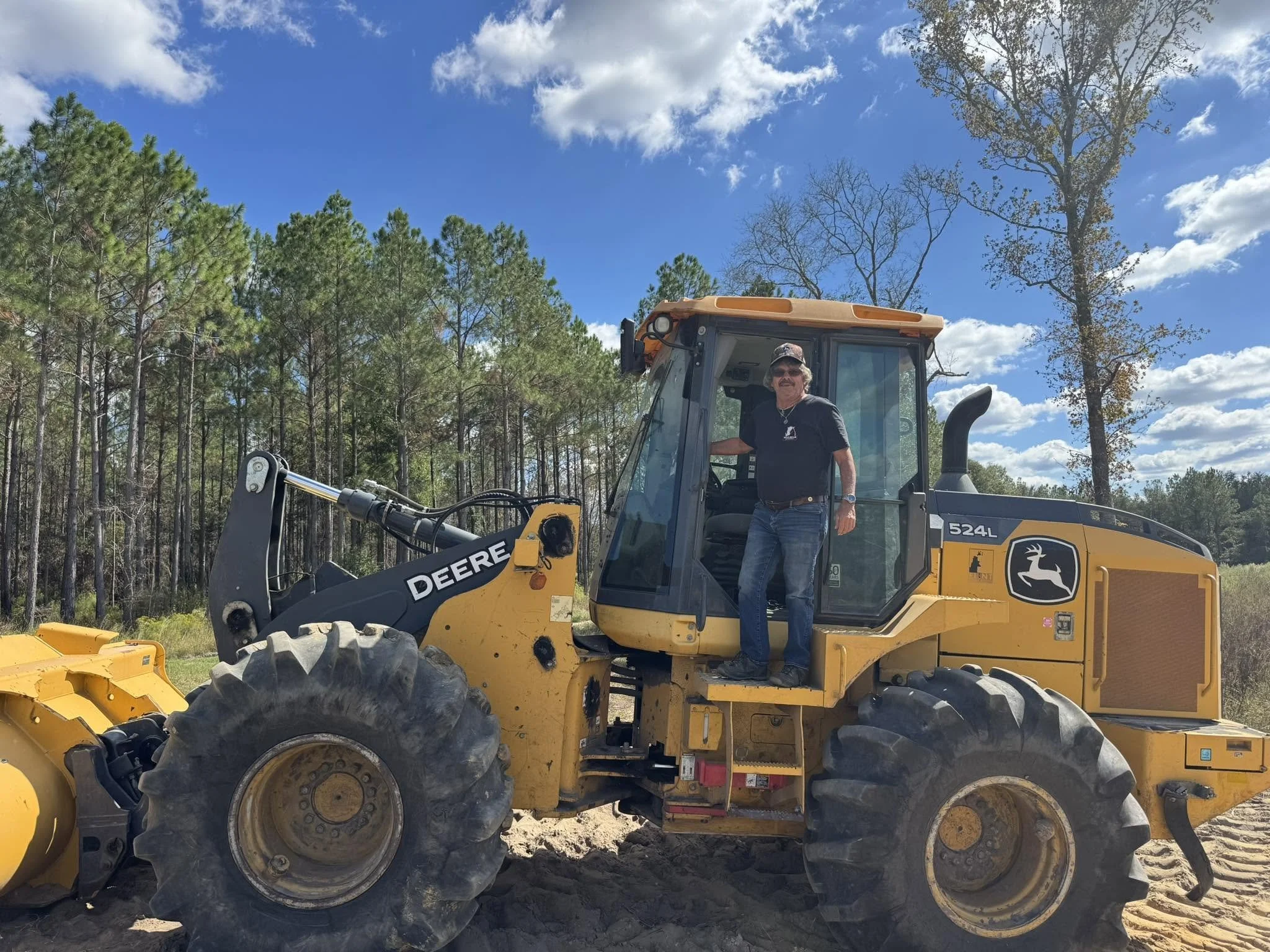 A man standing inside the cabin of a yellow John Deere construction vehicle, with a background of a forest and blue sky with clouds.