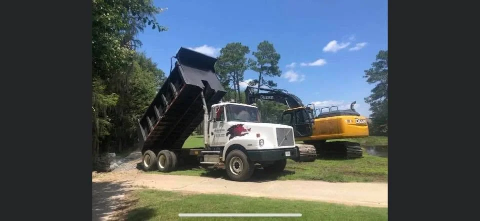 A white dump truck with a dragon decal on the side, a black excavator, and a yellow bulldozer parked on grass and dirt near trees and a river under a partly cloudy sky.