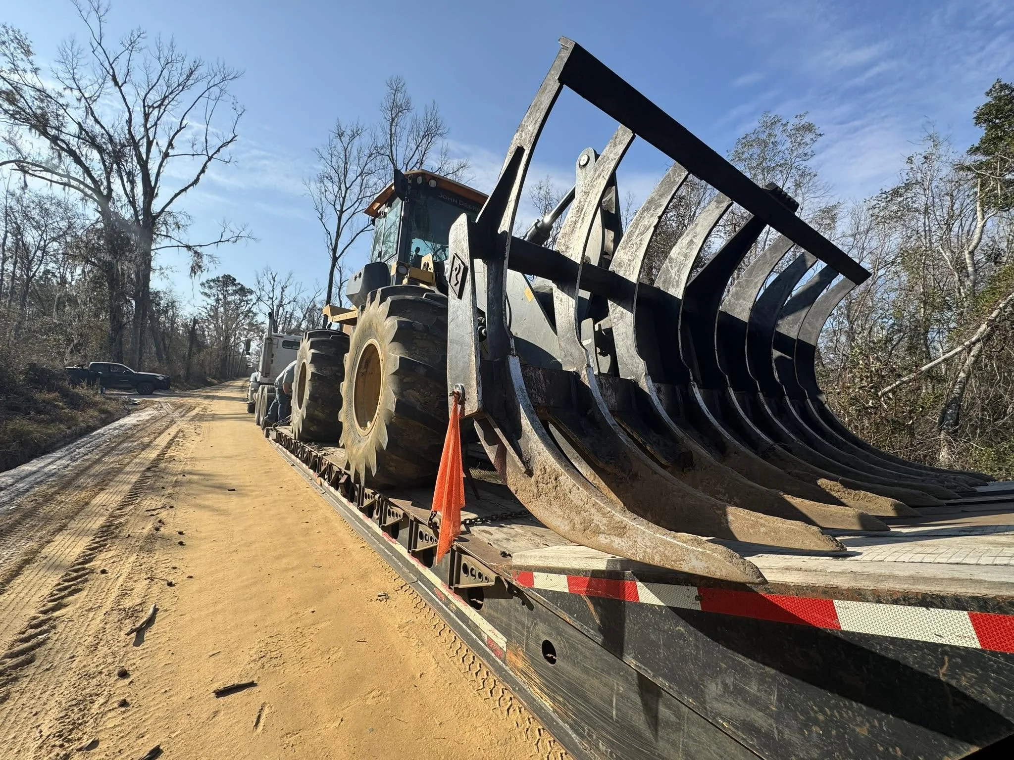 A large construction vehicle with a rake attachment on a flatbed trailer, parked on a dirt road surrounded by bare trees and a partly cloudy sky.