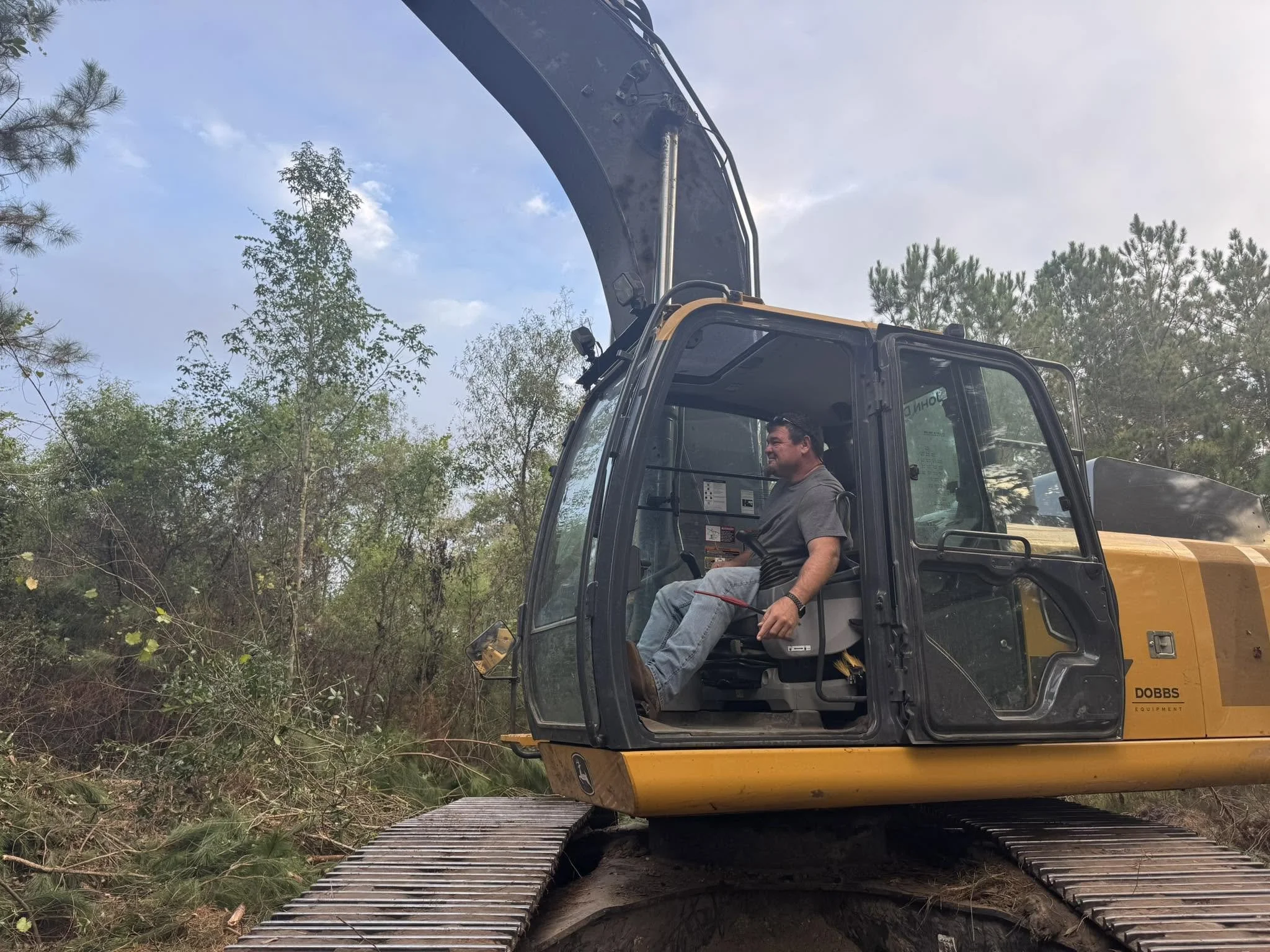 A man is riding in the cabin of a yellow excavator with a black arm, working in a wooded area with trees and bushes, and a sky with some clouds overhead.