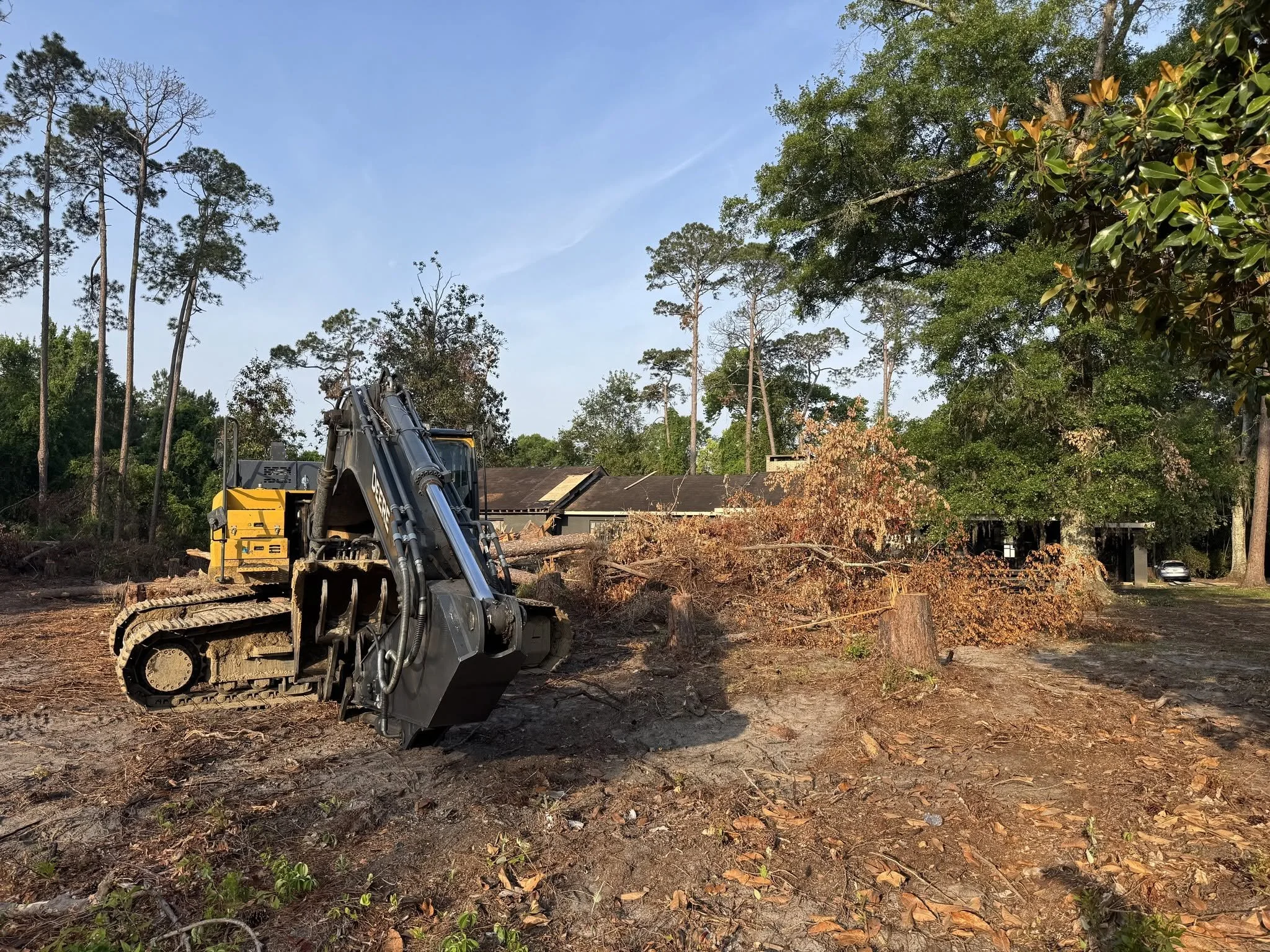 A construction site with a large excavator working on clearing land. There are trees and a house in the background under a clear blue sky.