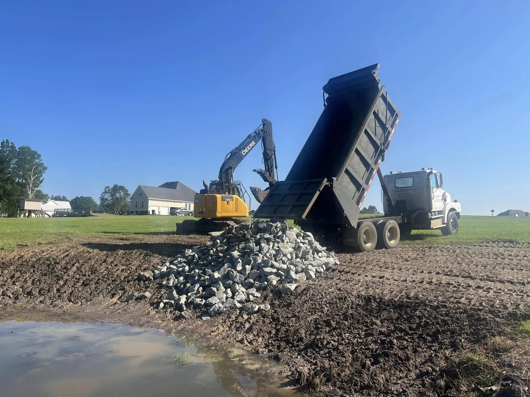 A construction site with a dump truck lifting its bed to unload rocks and soil, and an excavator nearby on a clear, sunny day, with houses and trees in the background.
