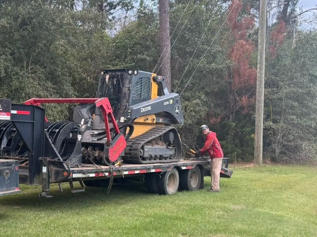 A man in a red jacket and beige pants standing next to a flatbed truck with a small John Deere construction vehicle on it, near a grassy area and power lines.