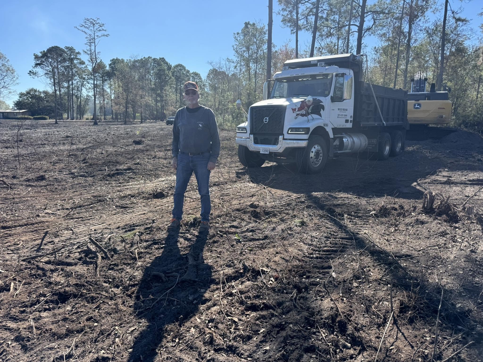A man wearing sunglasses and a cap standing near a bulldozer on freshly burned land with trees in the background.