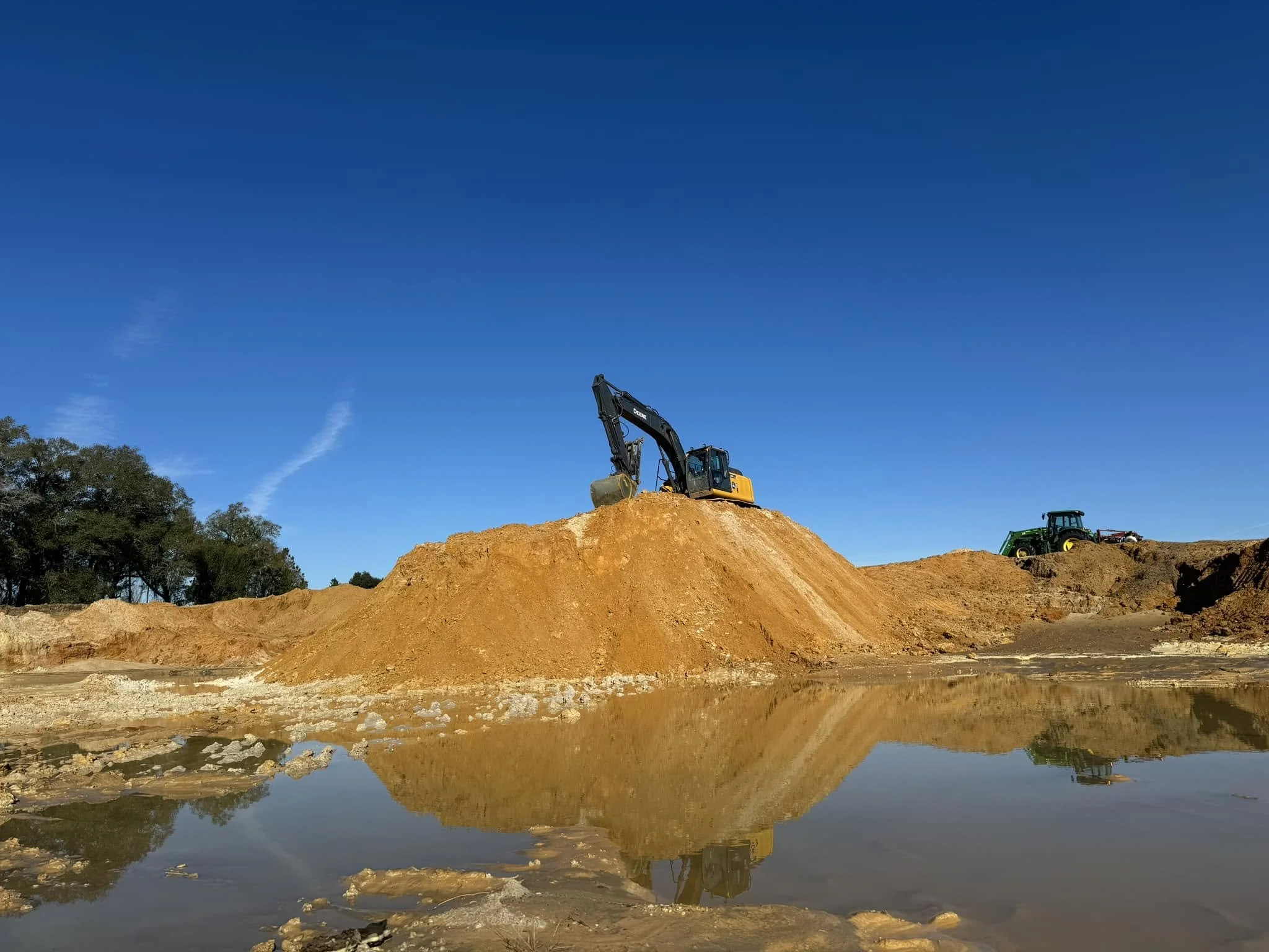 Construction site with a yellow excavator on a mound of dirt, a green tractor, and a body of water reflecting the scene, under a clear blue sky.