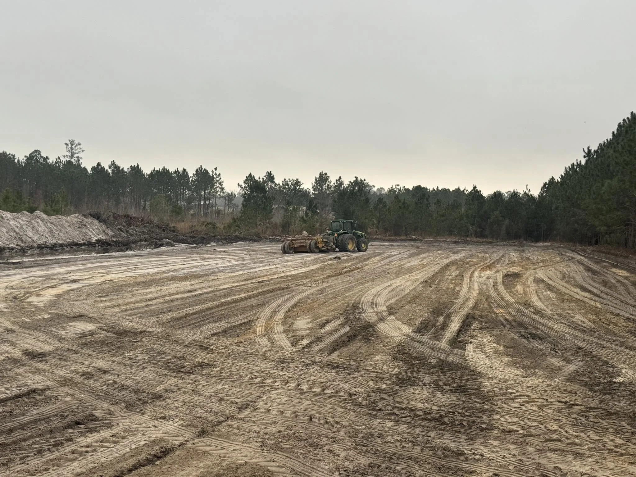 A tractor parks on a dirt construction site among trees with an overcast sky.