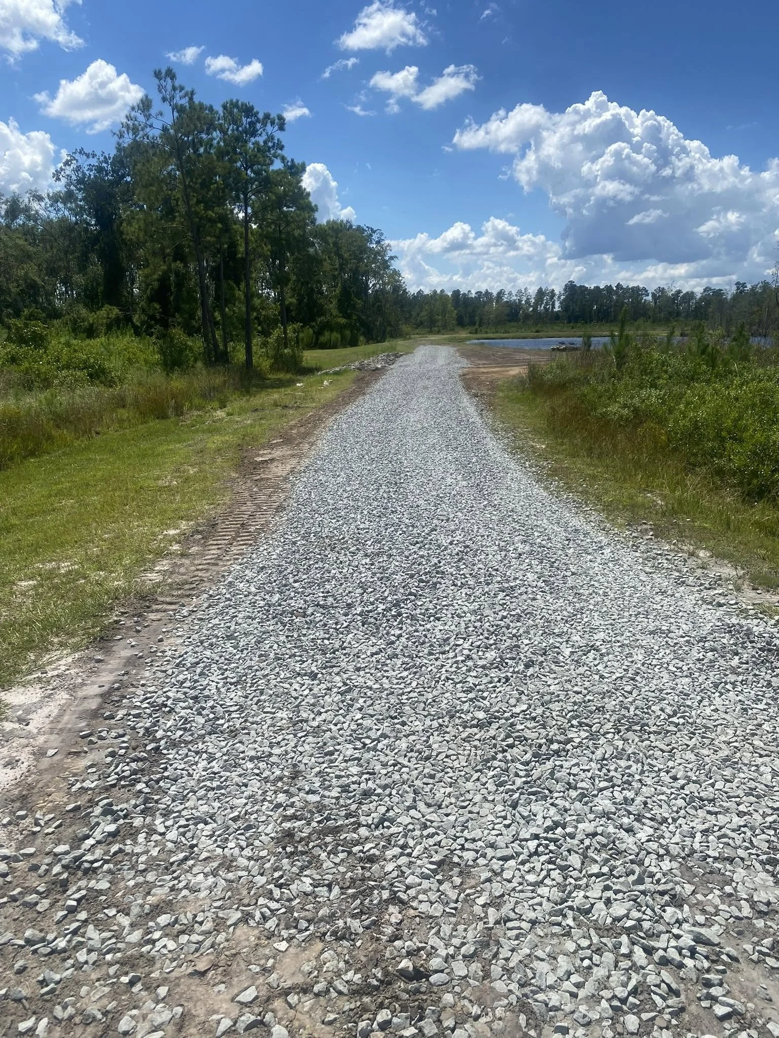 A gravel road runs through a natural landscape with green trees and bushes on either side, leading towards a body of water in the distance, under a blue sky with scattered clouds.