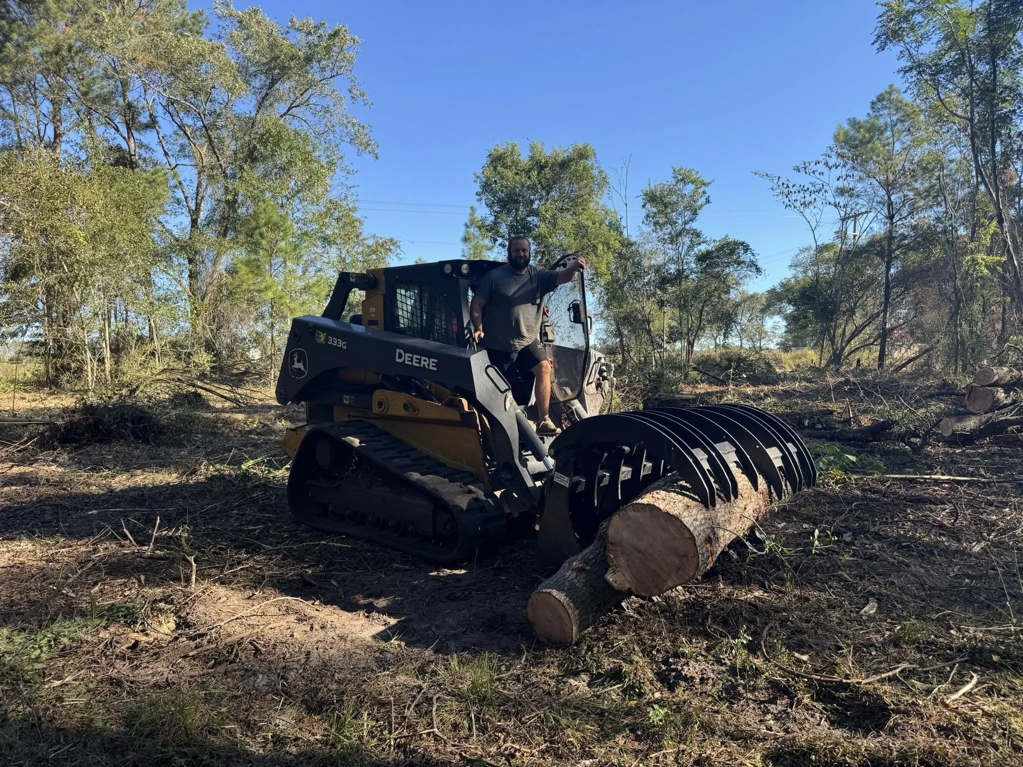 A man operating a John Deere compact tracked loader with a grapple attachment in a wooded area, lifting logs during a tree clearing or land management activity.