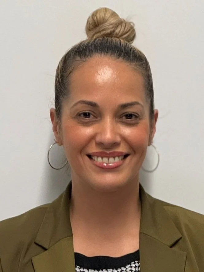 A woman with blonde hair styled in a high bun, smiling, wearing hoop earrings and a green jacket.