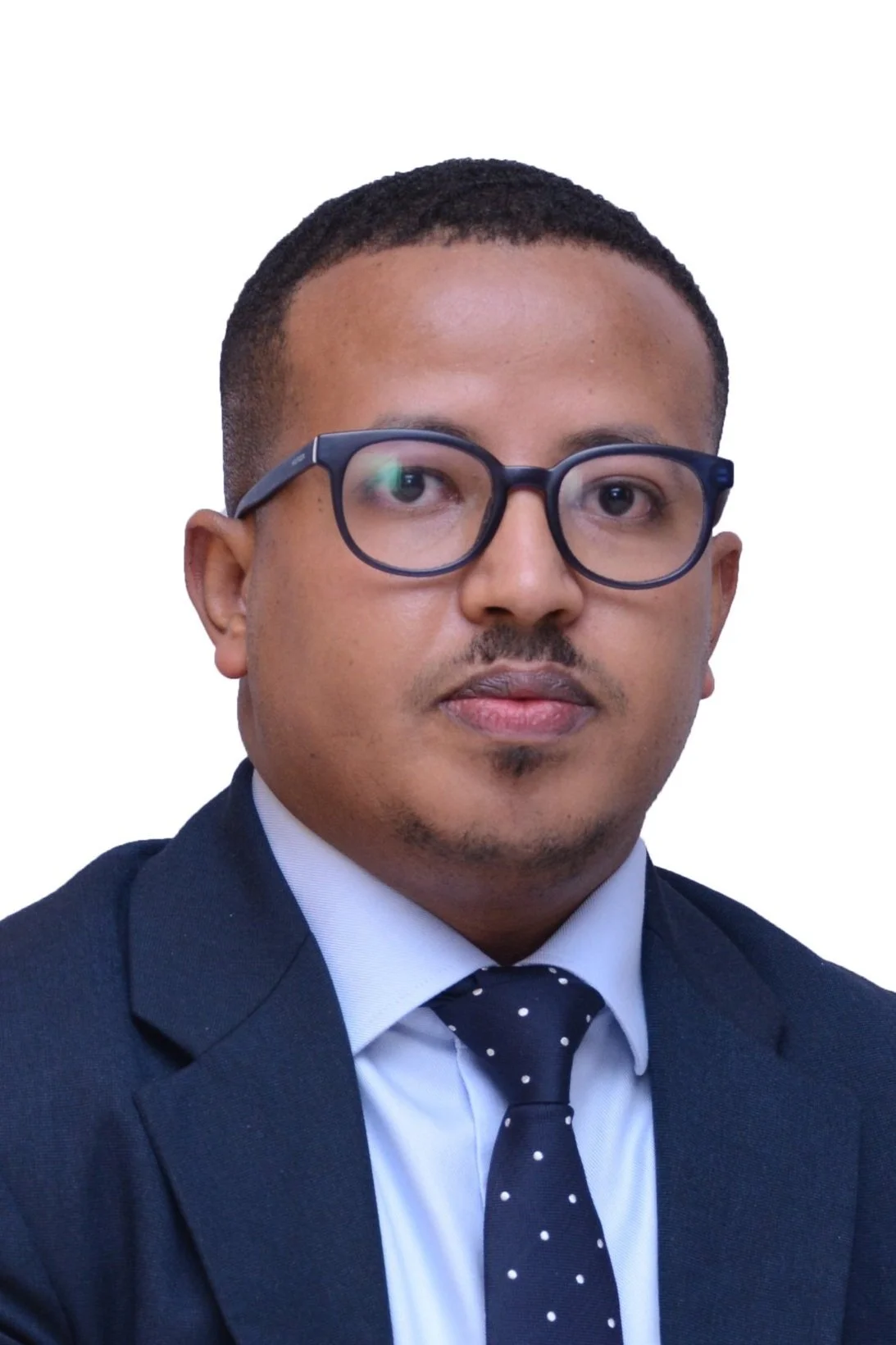 A man with short black hair and glasses wearing a navy blue suit, white shirt, and navy blue tie with white polka dots, against a plain white background.