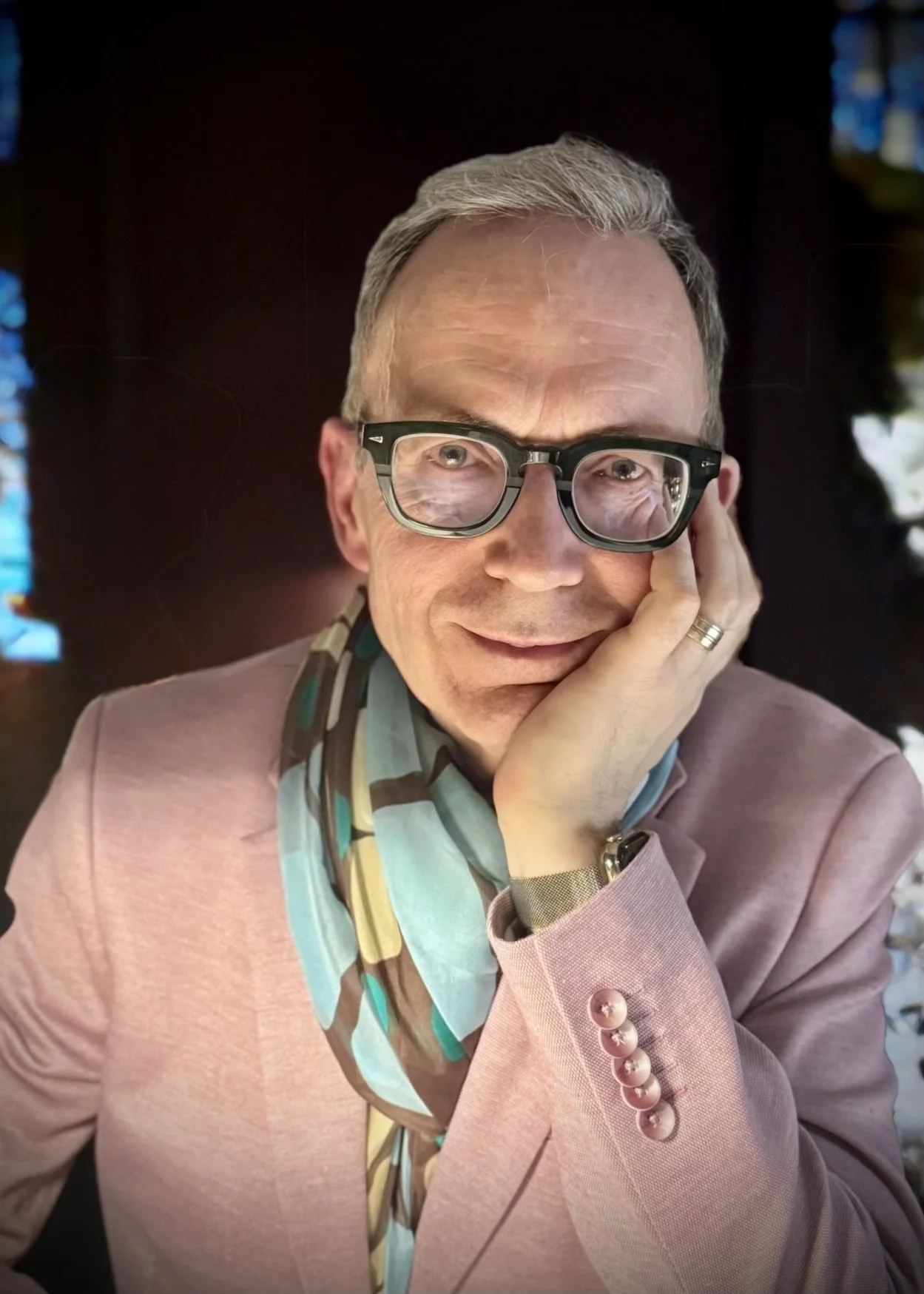 A smiling man with gray hair, glasses, and a pensive pose, resting his face on his hand, wearing a pink blazer and colorful scarf.