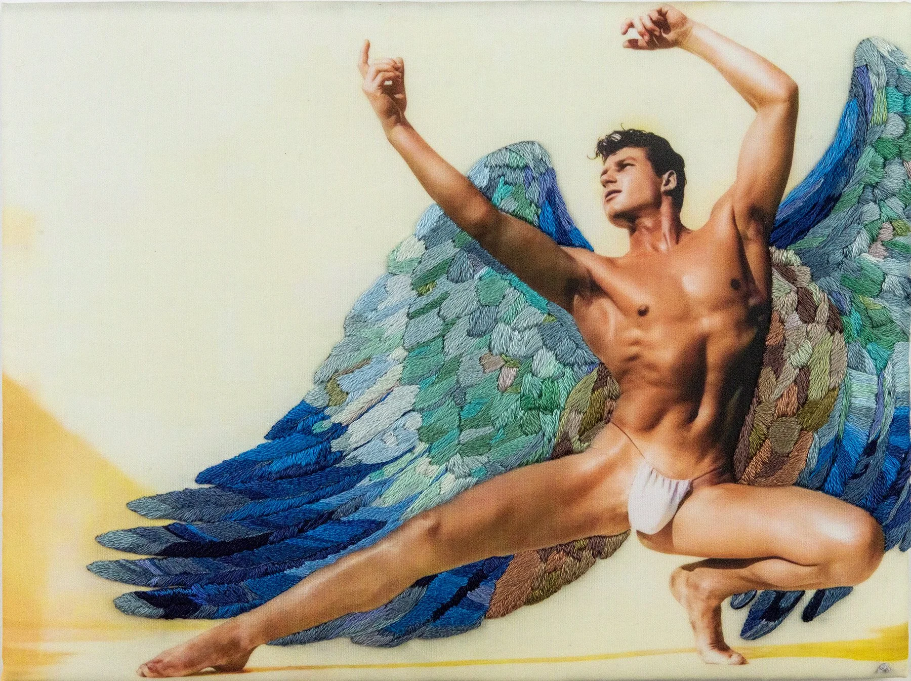 An artistic sculpture of a male figure with large, colorful feathered wings, posed with arms raised and head turned, set against a plain background.