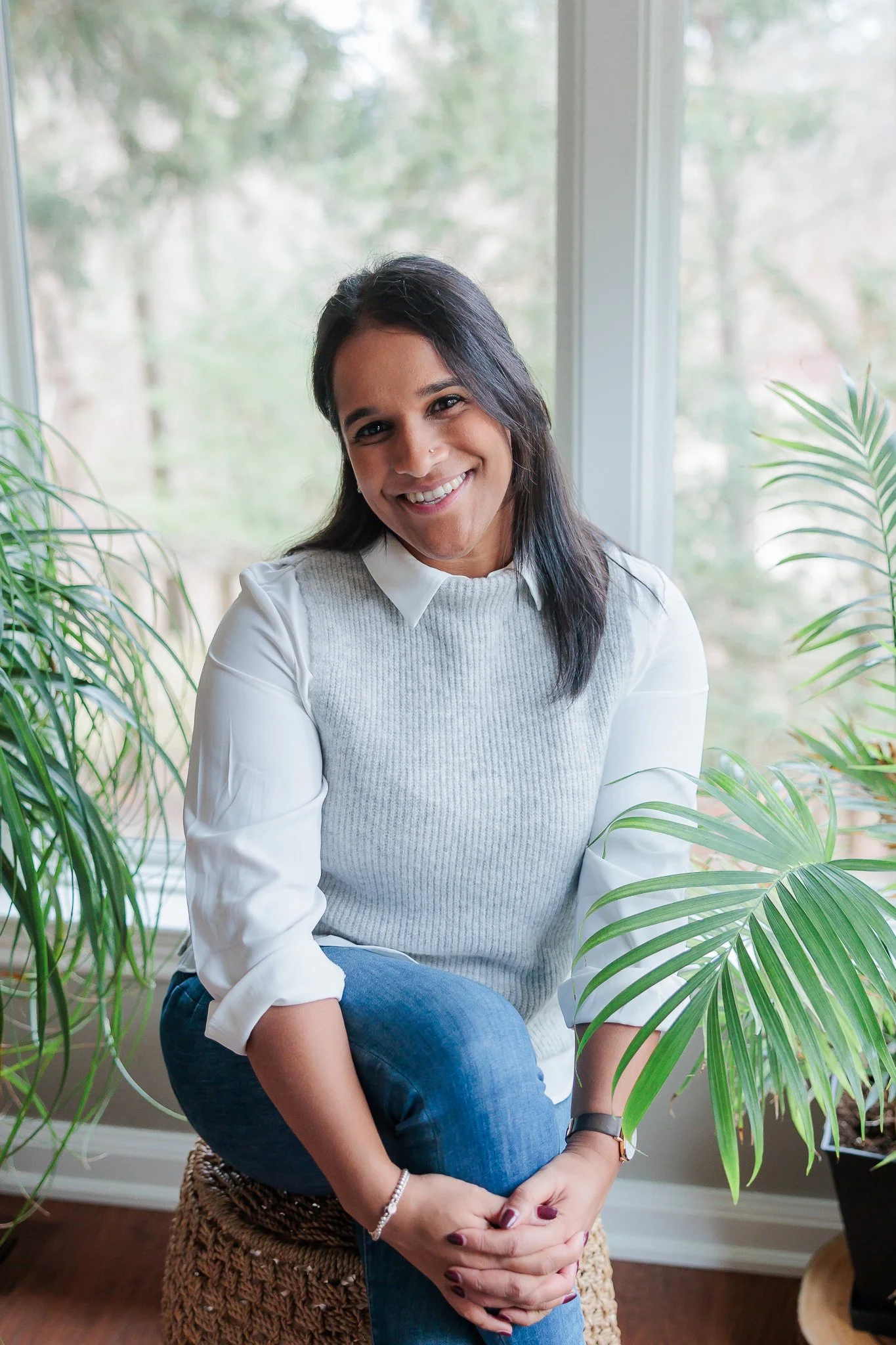 A woman smiling with dark hair wearing a white shirt, gray vest, and blue jeans, sitting near green houseplants by a window with trees outside.