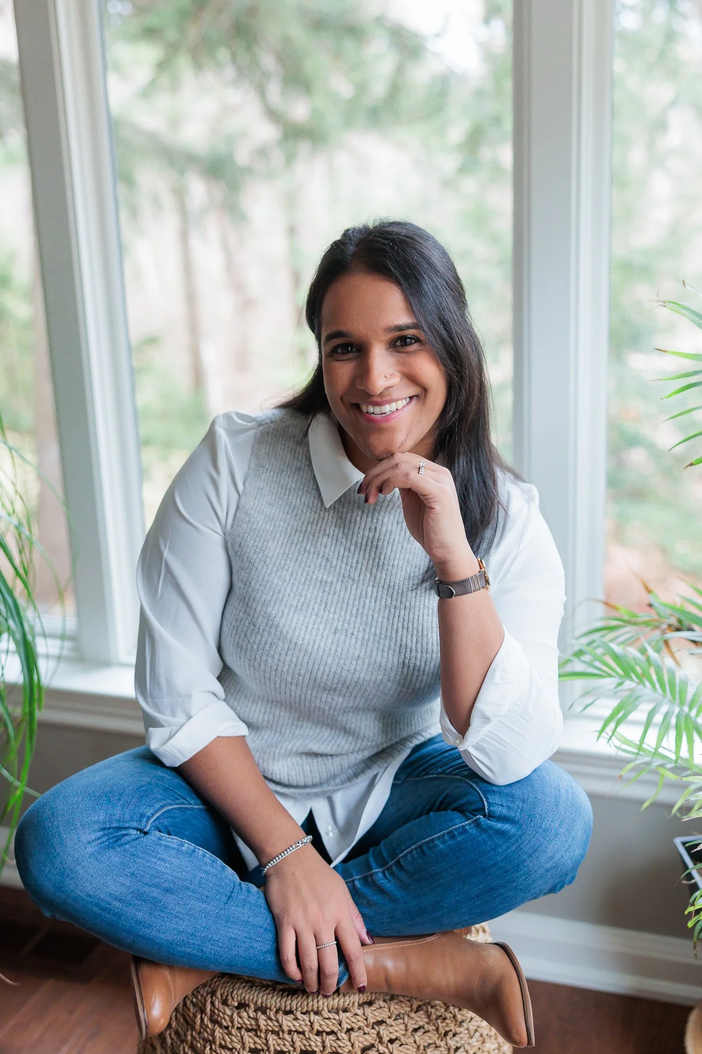 A woman sitting cross-legged on a woven ottoman, smiling while looking at the camera, with a window and green plants in the background.