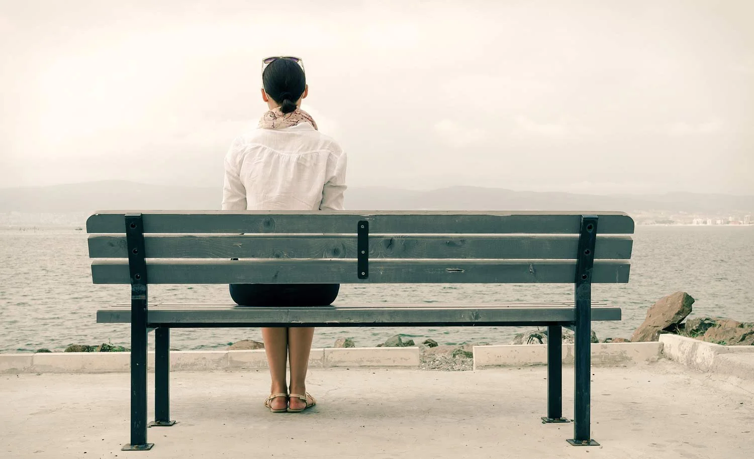 A woman with dark hair tied back, sitting on a wooden bench by the water, looking out at the ocean and distant mountains, wearing a white shirt, a scarf, and sandals.