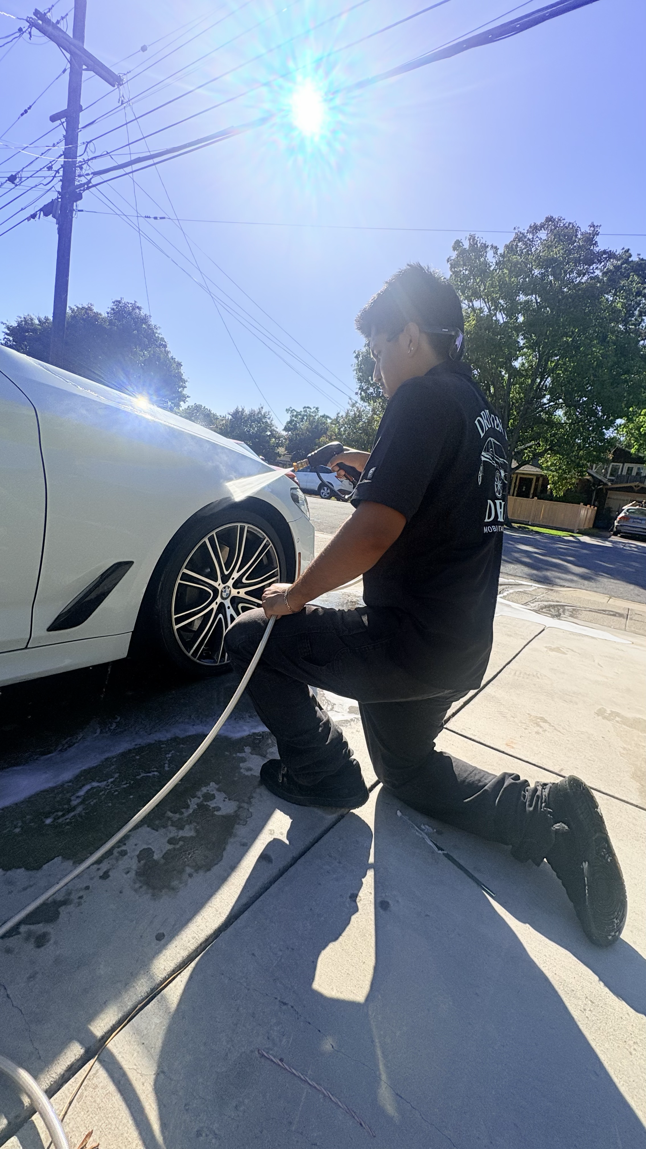 A person washing a white car with a hose outside on a sunny day, kneeling on one knee on the driveway.