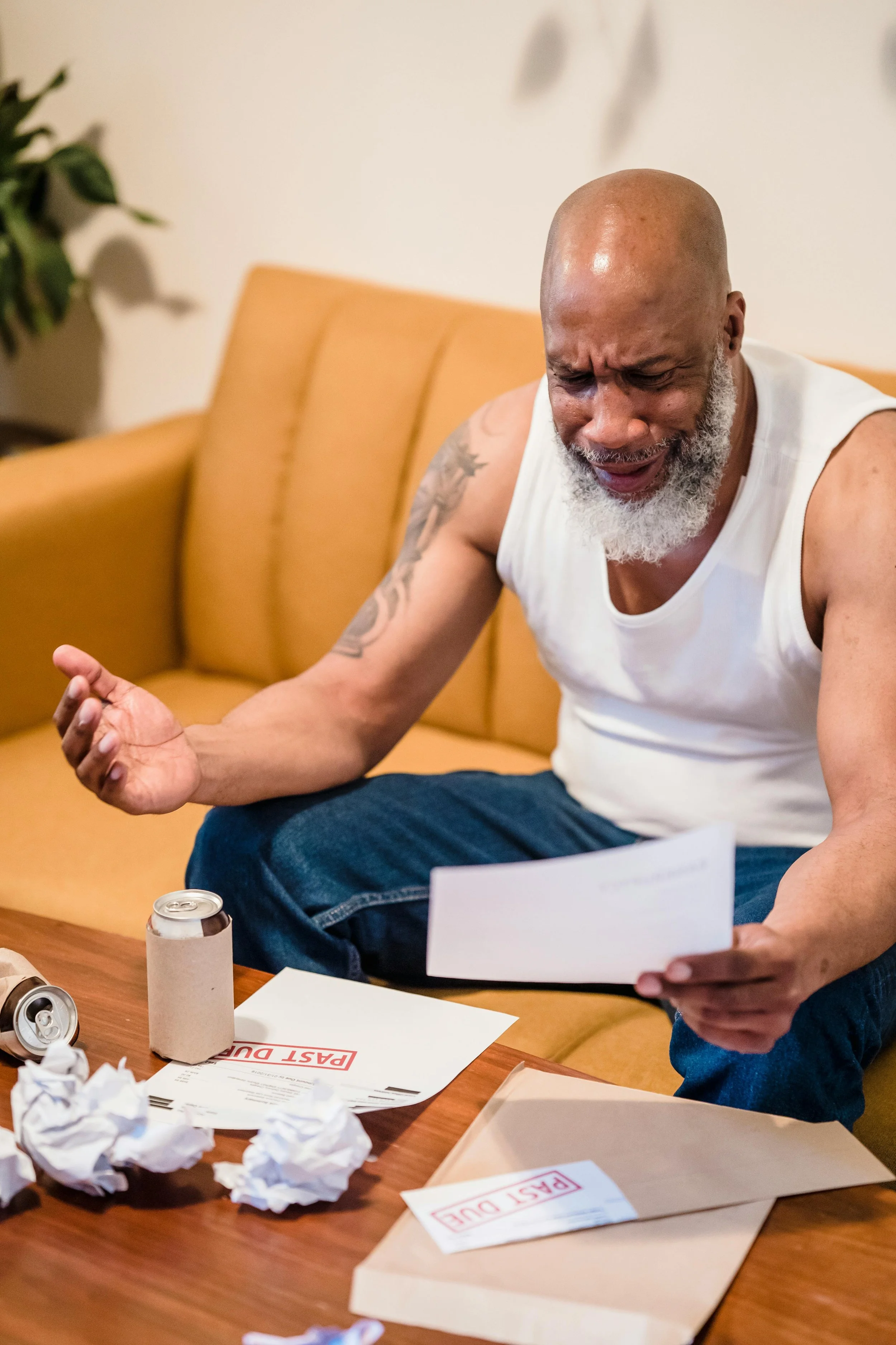 An older man with a bald head and gray beard is sitting on a yellow couch, looking distressed and crying while holding a piece of paper. In front of him on the table are crumpled tissues, a couple of crushed cans, and several white paper receipts or documents, some with the words 'PAST DUE' in red.
