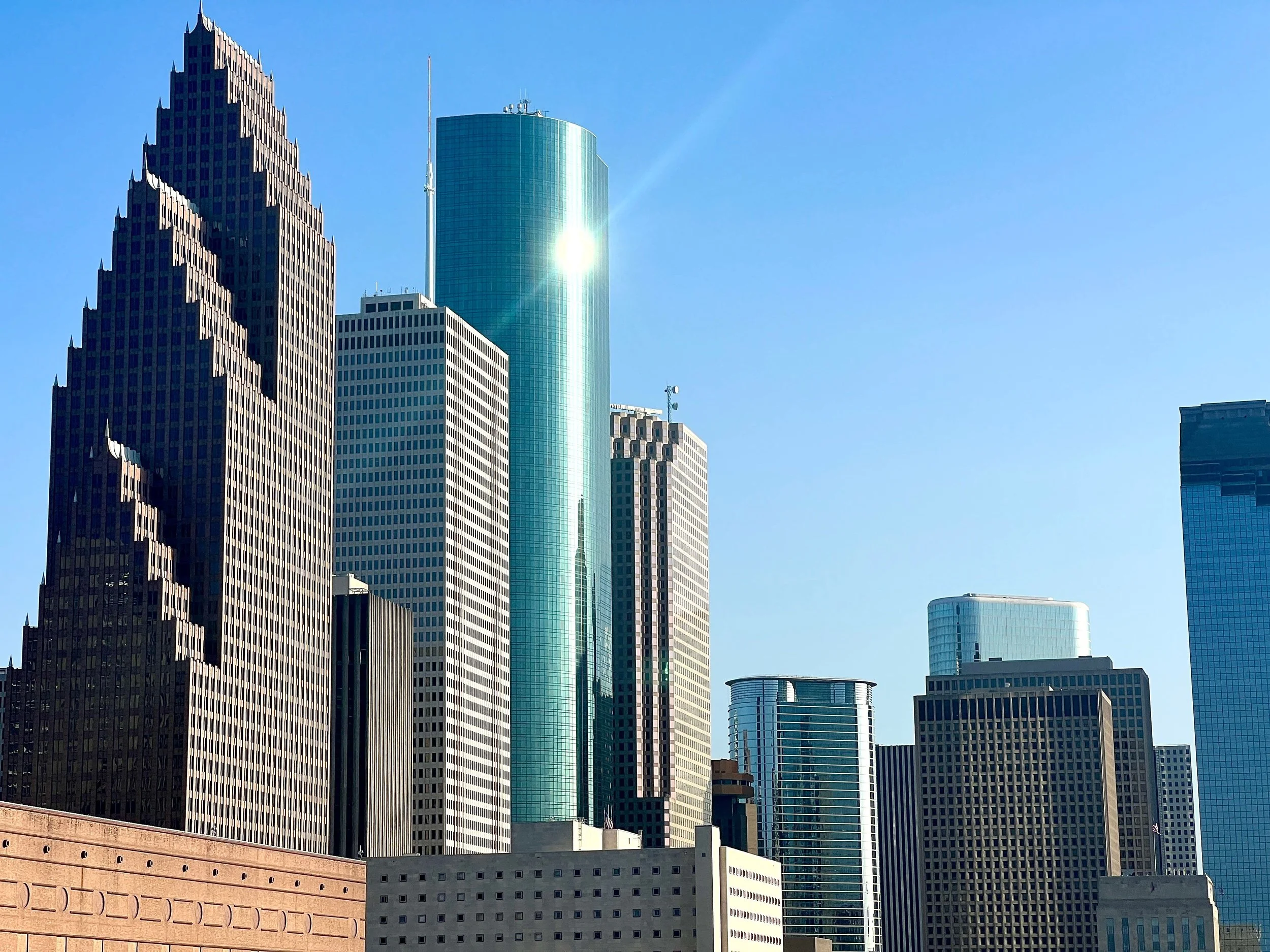 Skyline of several tall modern skyscrapers with reflective glass facades, with the sun shining off one building, against a clear blue sky.
