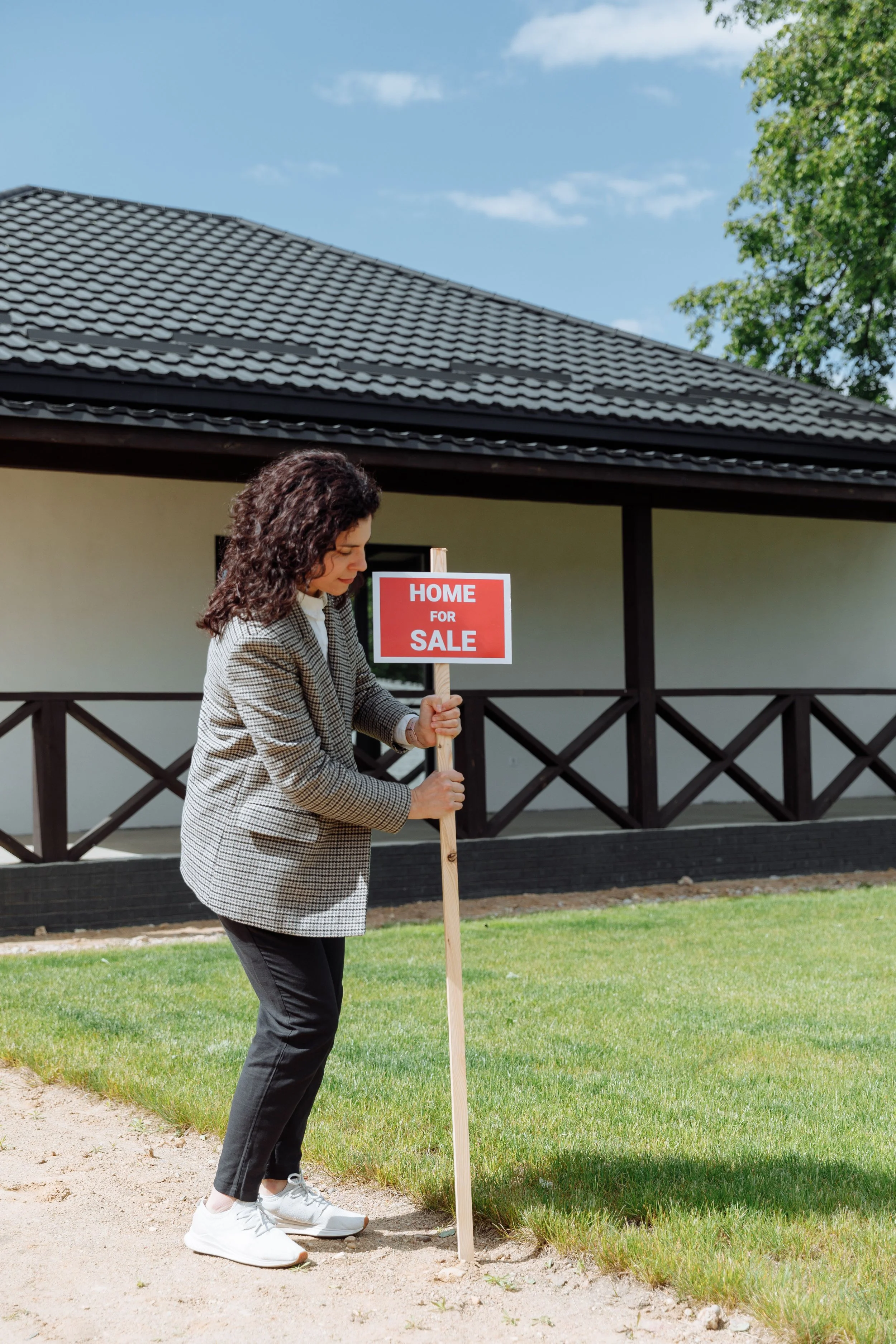 Woman holding a 'Home for Sale' sign, standing on a lawn outside a house with a dark tiled roof and a tree in the background.