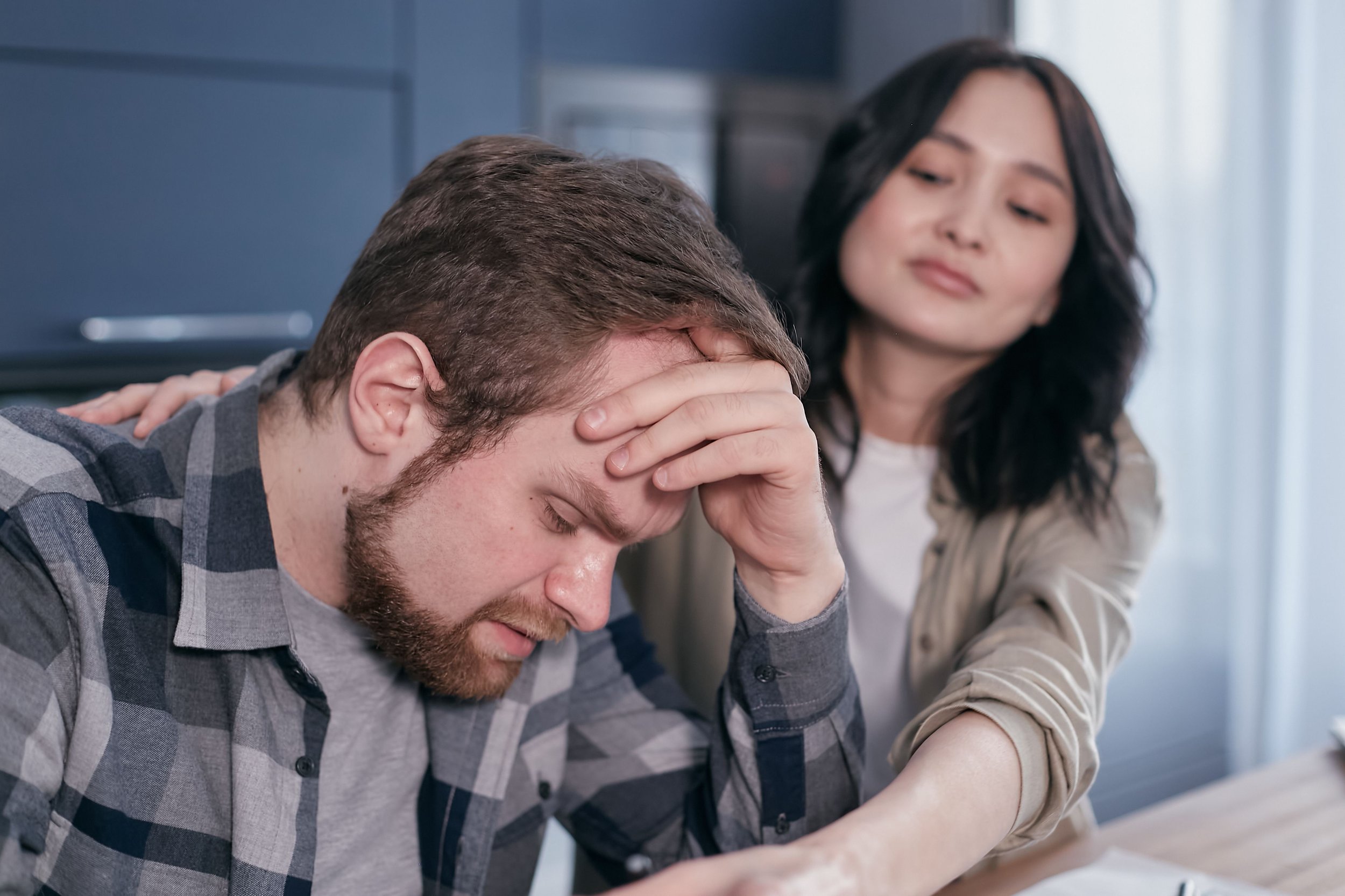 A distressed man with a beard, head in hand, sitting at a table with a woman beside him, appearing concerned and comforting.