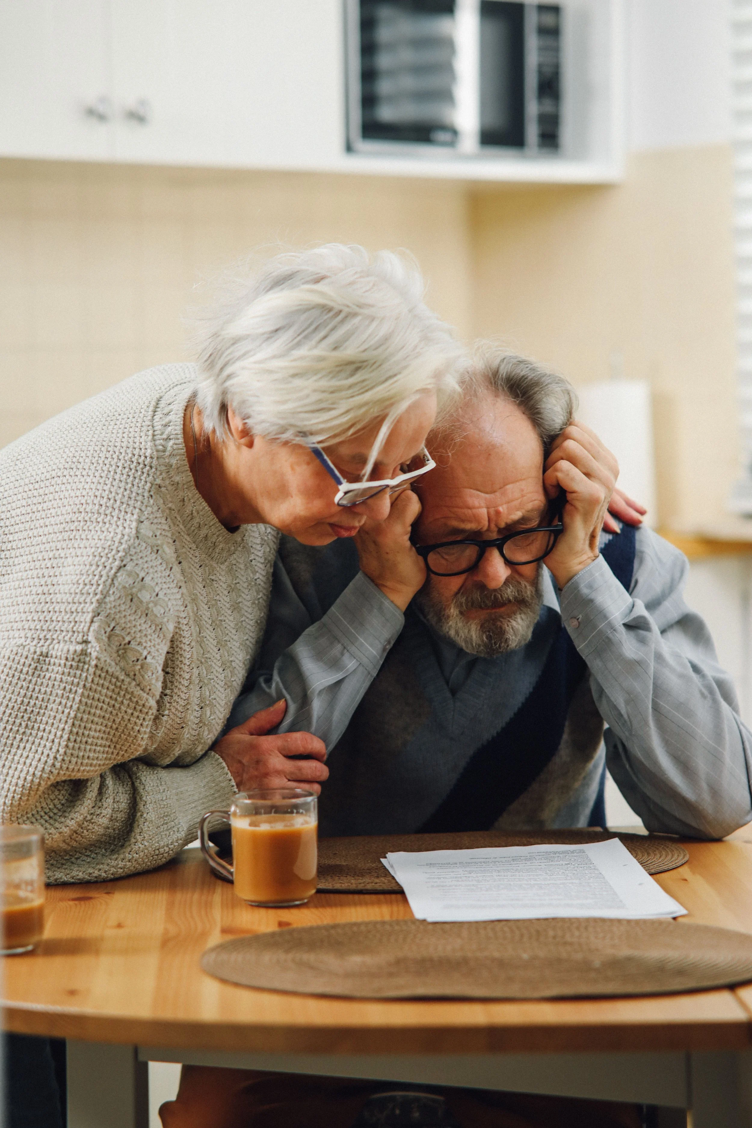 An elderly man with glasses and a beard, sitting at a wooden table, is leaning over and covering his ears with both hands, appearing distressed. An elderly woman with white hair and glasses is standing next to him, leaning over him, and appears to be comforting or speaking to him. There are two glasses of what looks like coffee or tea on the table, and some papers are spread out in front of the man.