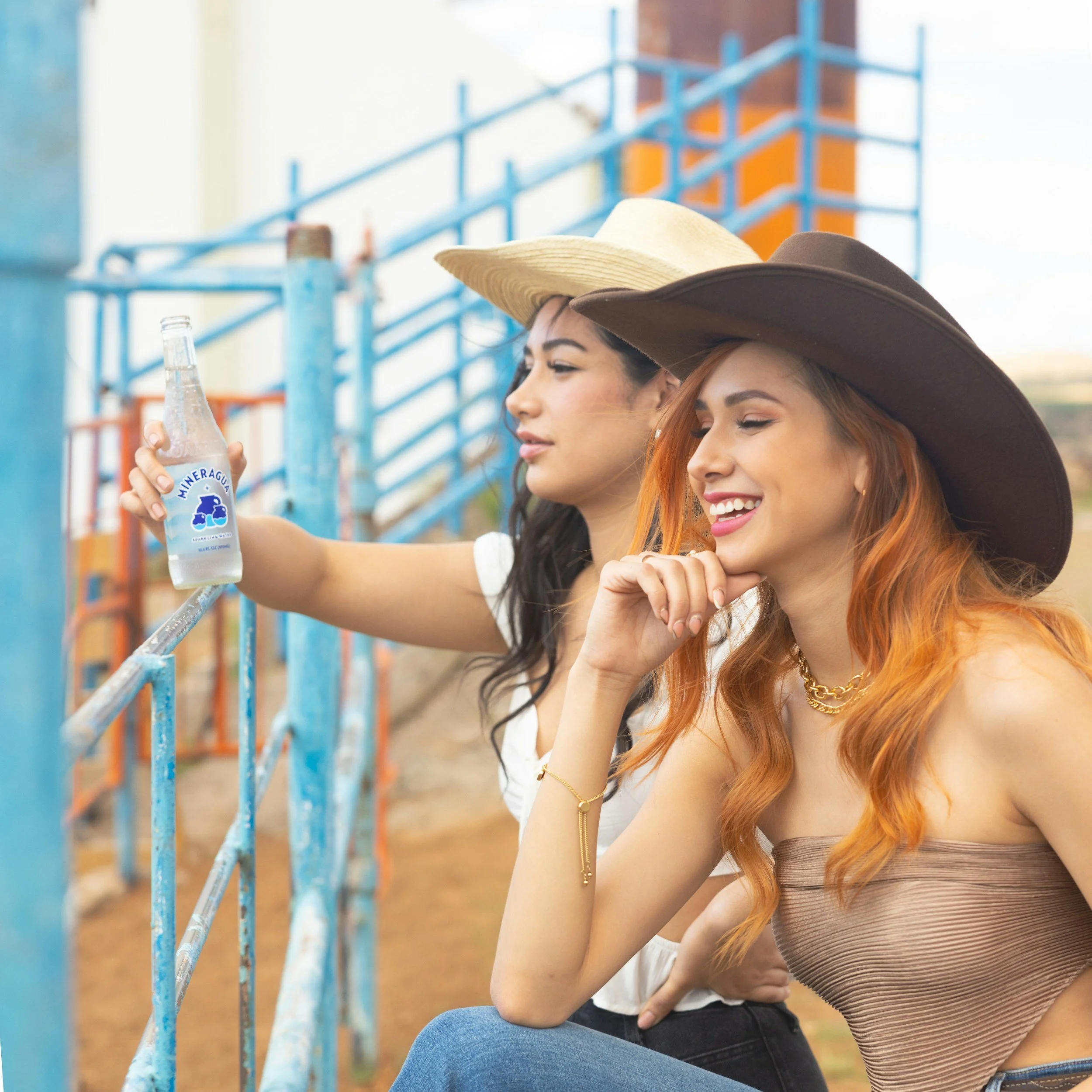 Two women with wide-brimmed hats sitting outdoors near a blue metal fence, with a blurred playground in the background. One woman is holding a water bottle labeled 'Mineragua' and smiling.