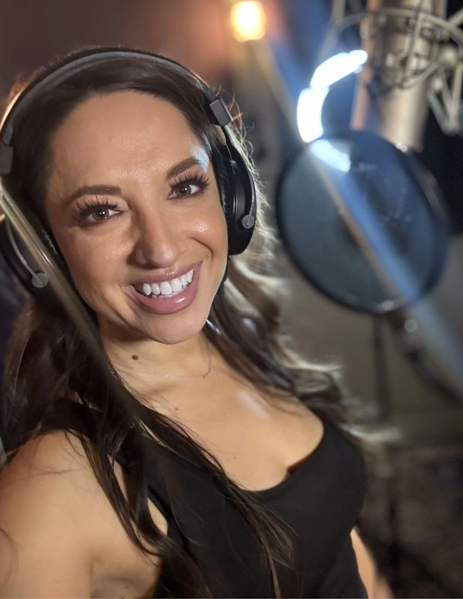 A woman smiling with long dark hair, wearing a headset, in a recording studio with microphones and bright lights in the background.