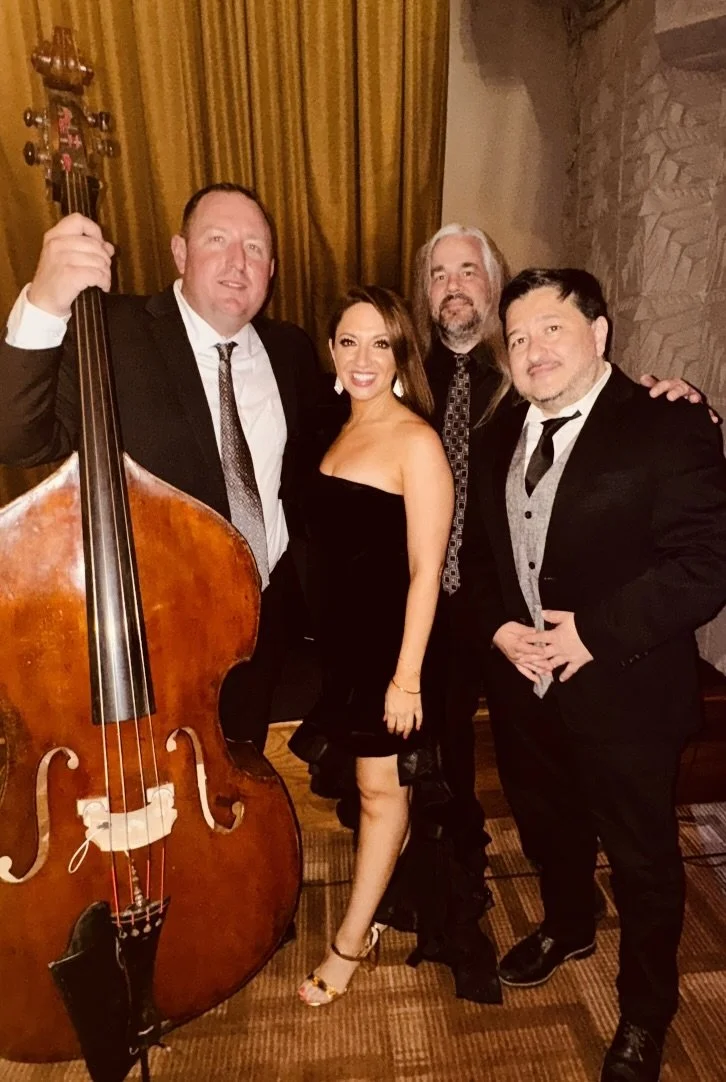Four people dressed in formal attire, standing together indoors with a large double bass and a background of curtains and stone wall.