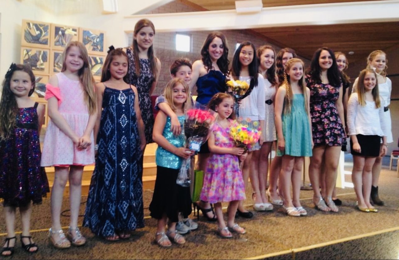 Children and women dressed in dresses standing on a stage, some holding bouquets of flowers, in a church or community hall setting.