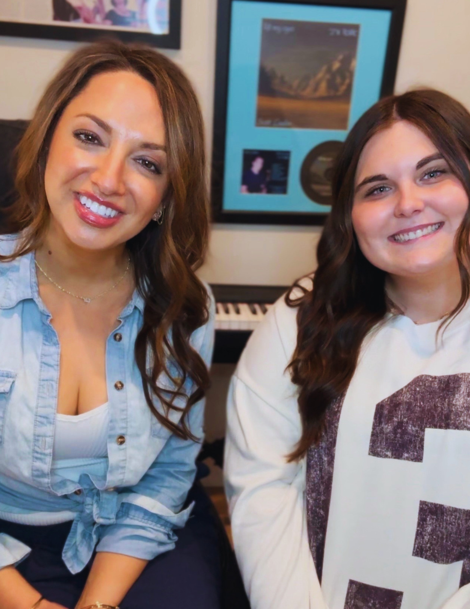 Smiling woman with light brown hair wearing a denim shirt and white top, sitting next to another smiling woman with dark brown hair, in front of a framed picture of mountains on the wall.