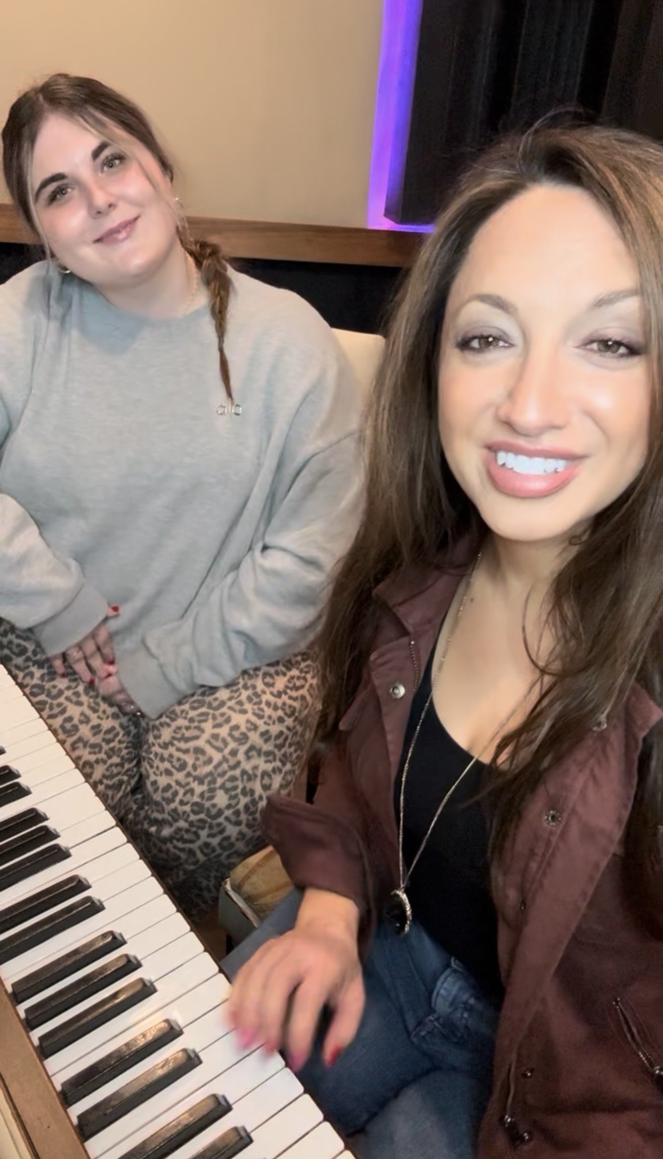 Two women sitting at a piano, one playing the keys and the other smiling at the camera.