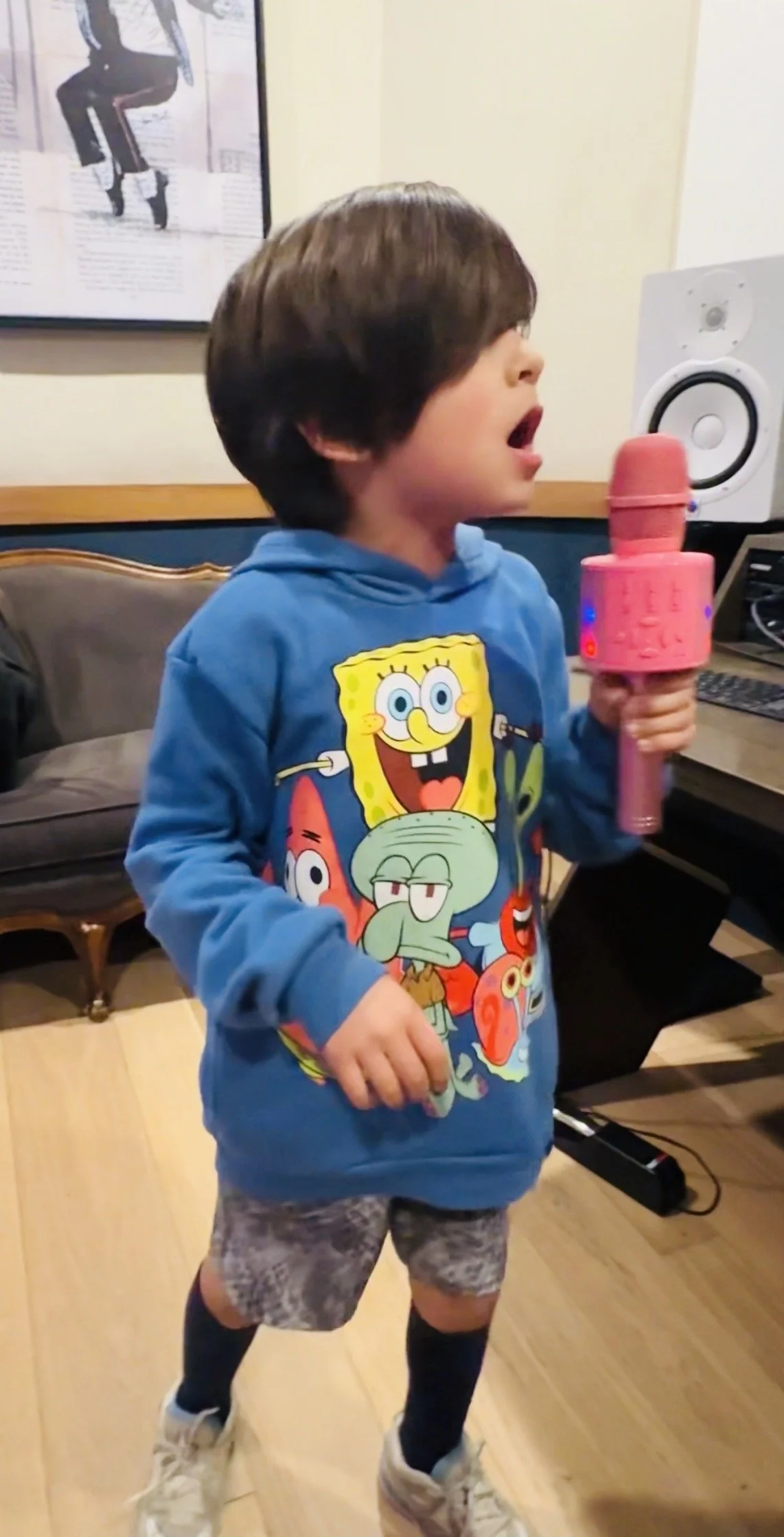 A young boy singing into a pink microphone in a room with a keyboard, speaker, and framed picture on the wall. He is wearing a blue SpongeBob SquarePants hoodie, gray shorts, and sneakers.