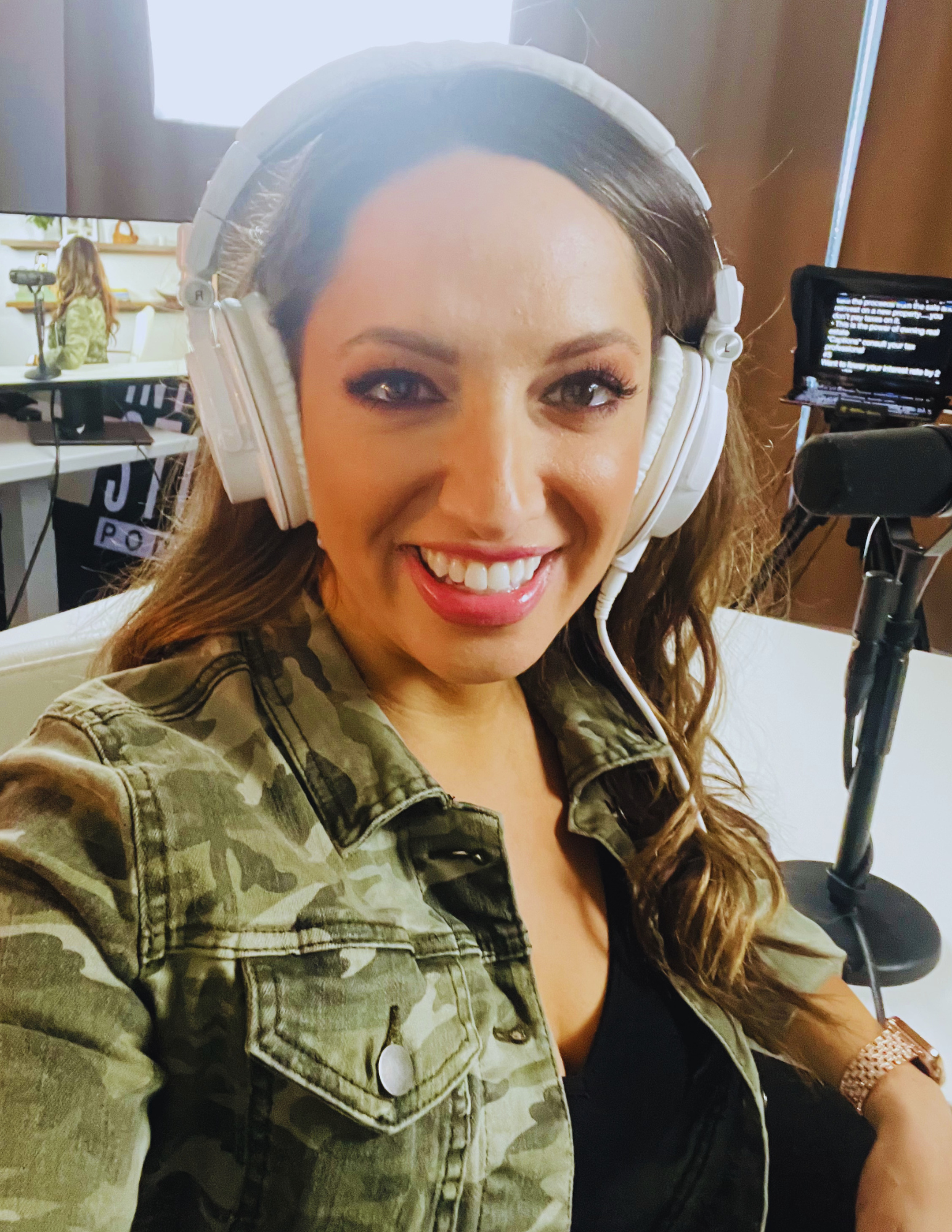 A woman wearing camouflage jacket with large white headphones smiling at the camera, with a social media monitor on a stand beside her.