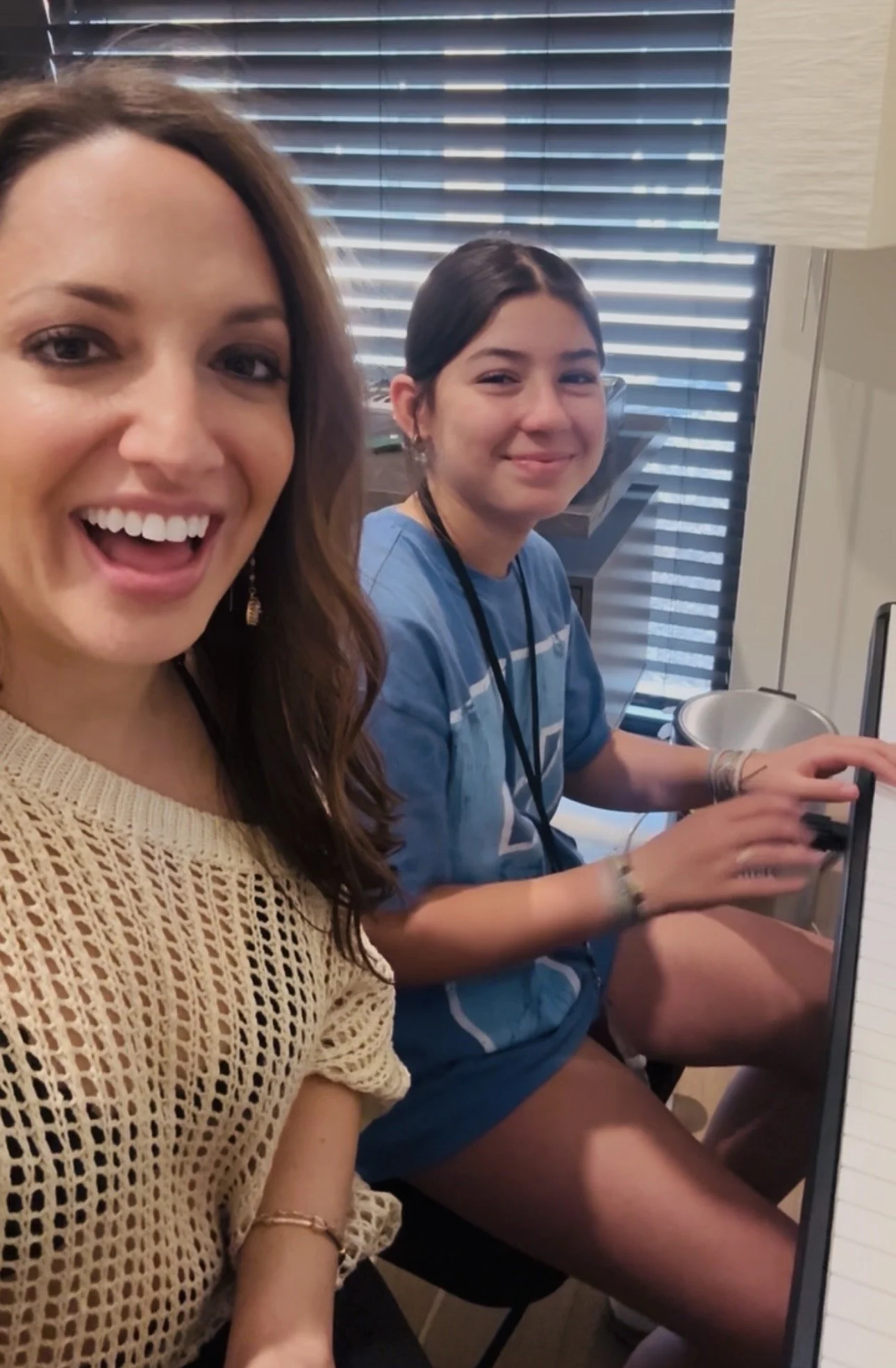 Two women taking a selfie in front of a computer. One woman has brown hair and is smiling with her mouth open, wearing a beige crochet top. The other woman has dark hair, is smiling, and is wearing a blue shirt, sitting at a desk with a computer mouse in her hand.