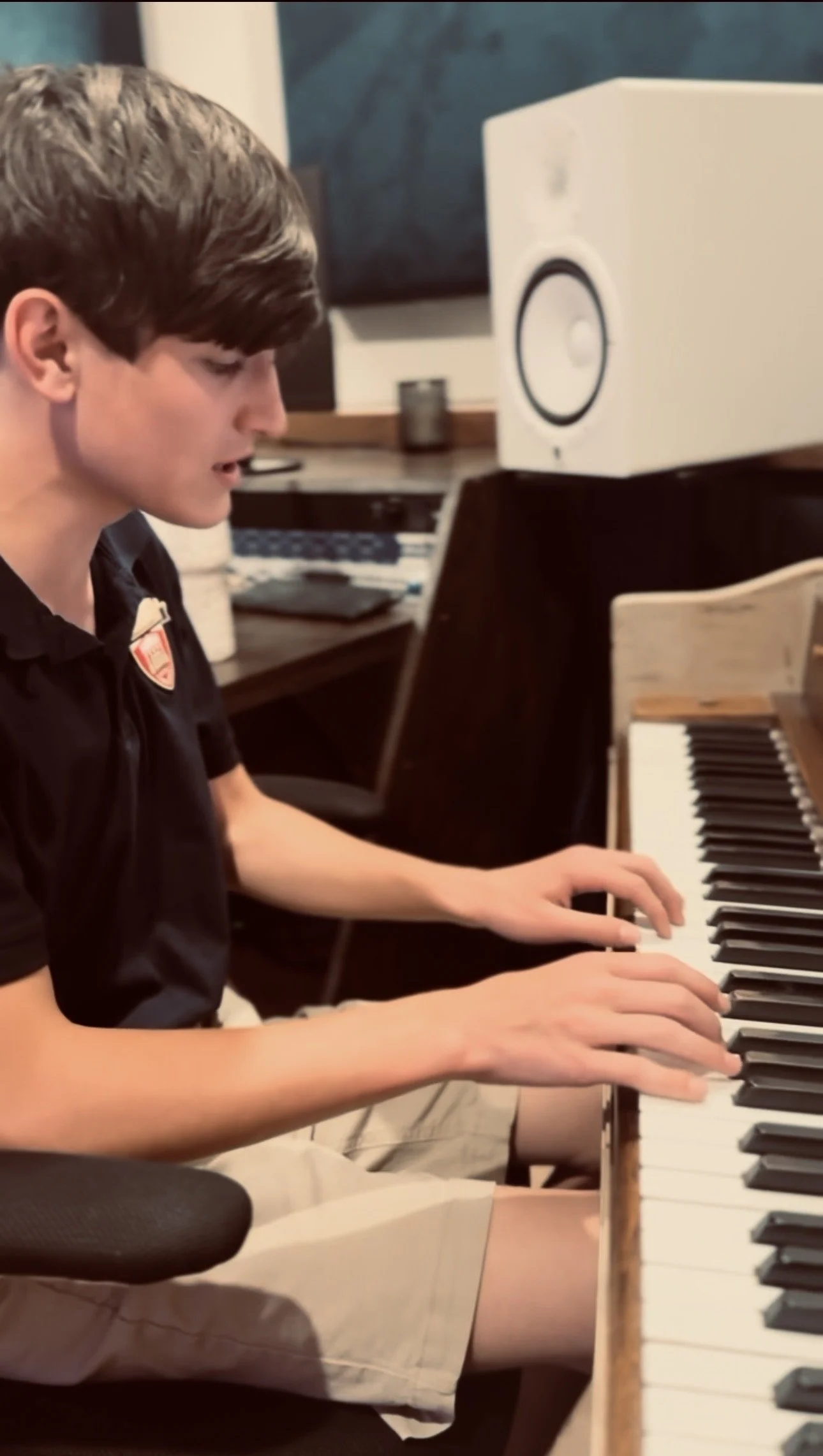 A young person playing piano, viewed from the side, in a room with computer and speaker equipment visible in the background.