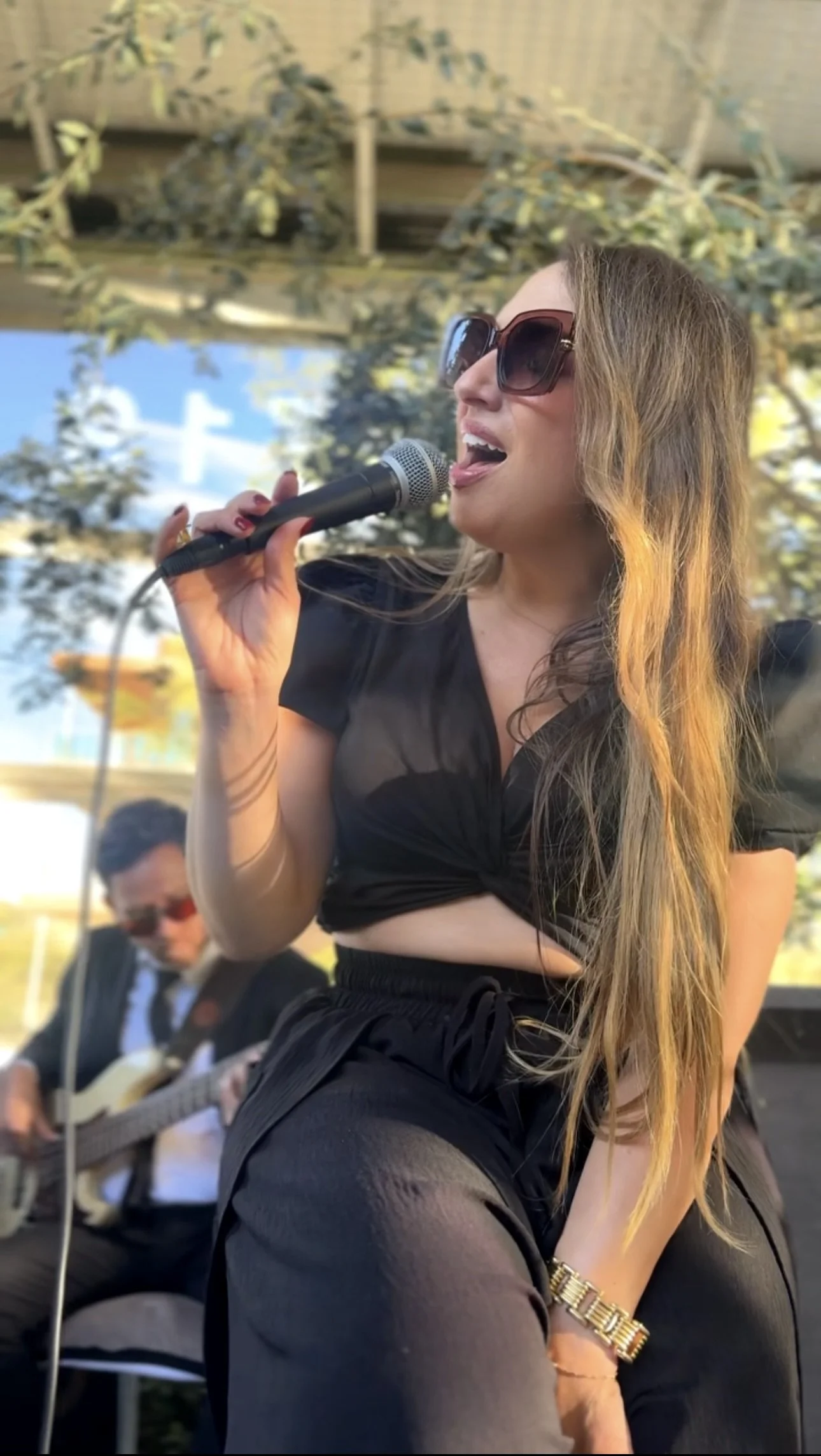 A woman singing into a microphone with sunglasses, wearing a black crop top and black pants, with long hair and a gold watch, outdoors with trees and a blue sky in the background. A man playing guitar is visible in the background.